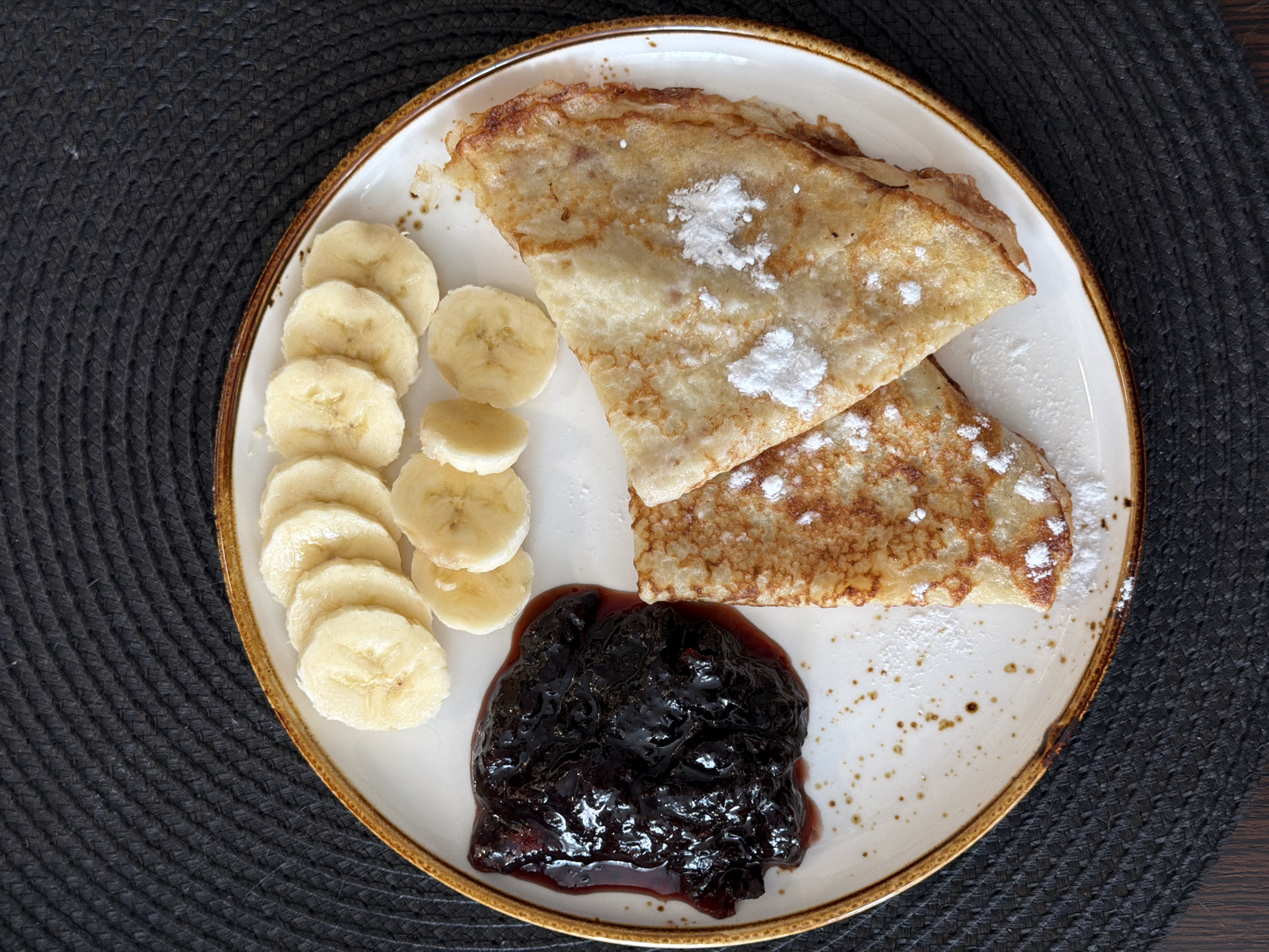 The image shows a plate of food on a dark-colored surface. On the plate are two folded crepes dusted with powdered sugar, a pile of sliced bananas, and a dollop of dark jam. The plate is round, white with a brown rim. The background is a woven dark material. No evidence suggests this picture was taken in Засечное, Russia.