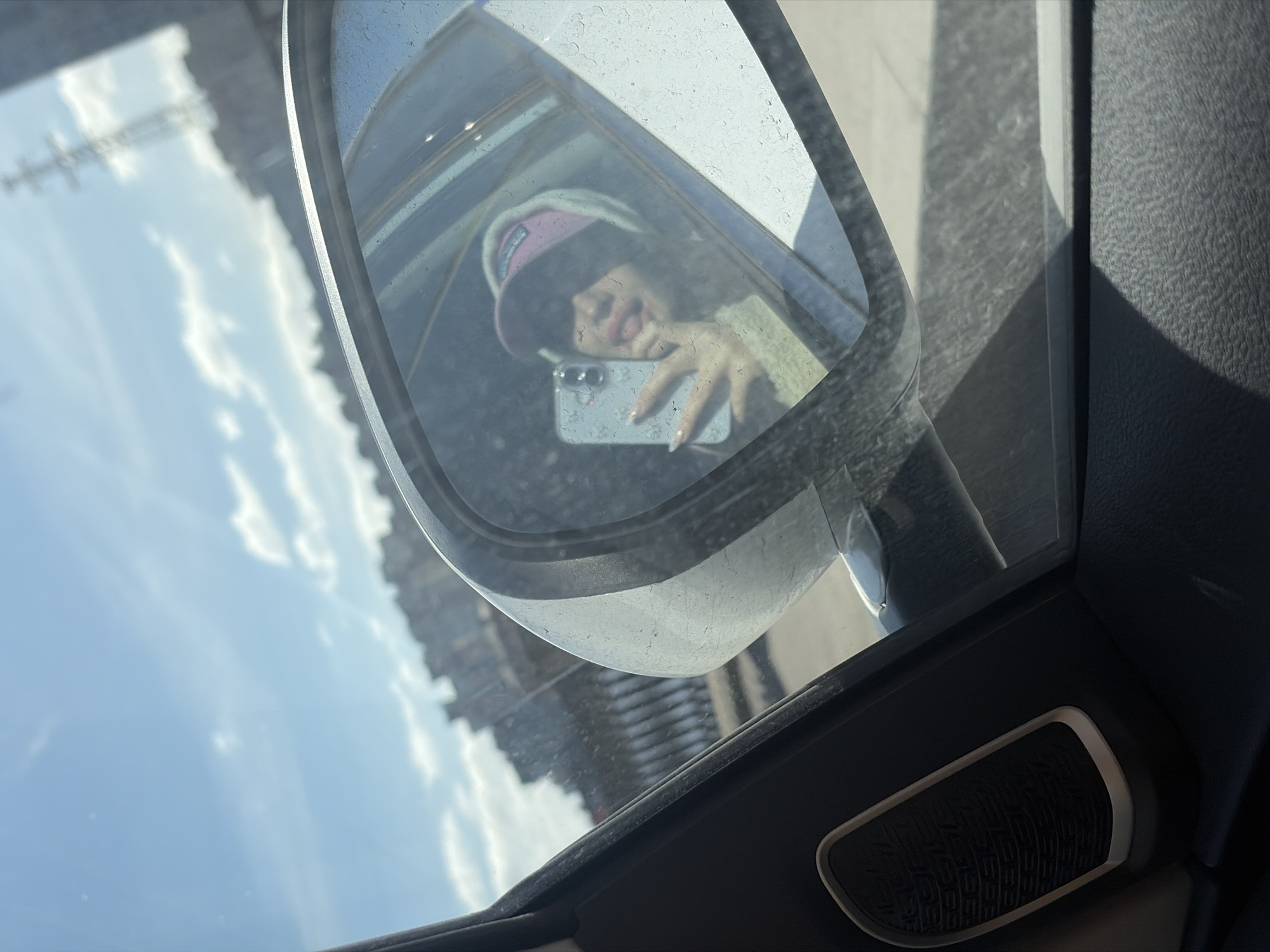 A selfie is taken in the driver's side mirror of a car, reflecting a person wearing a pink and white hat, a light-colored sweater, and holding a smartphone. The reflection shows the person sticking their tongue out. The car is parked or stopped, with the view outside the window showing a sky with clouds and a building structure resembling power lines or a transmission tower. The interior of the car is visible with a dark panel and a speaker. The exact location in St. Petersburg, Russia is not discernible based solely on this image.