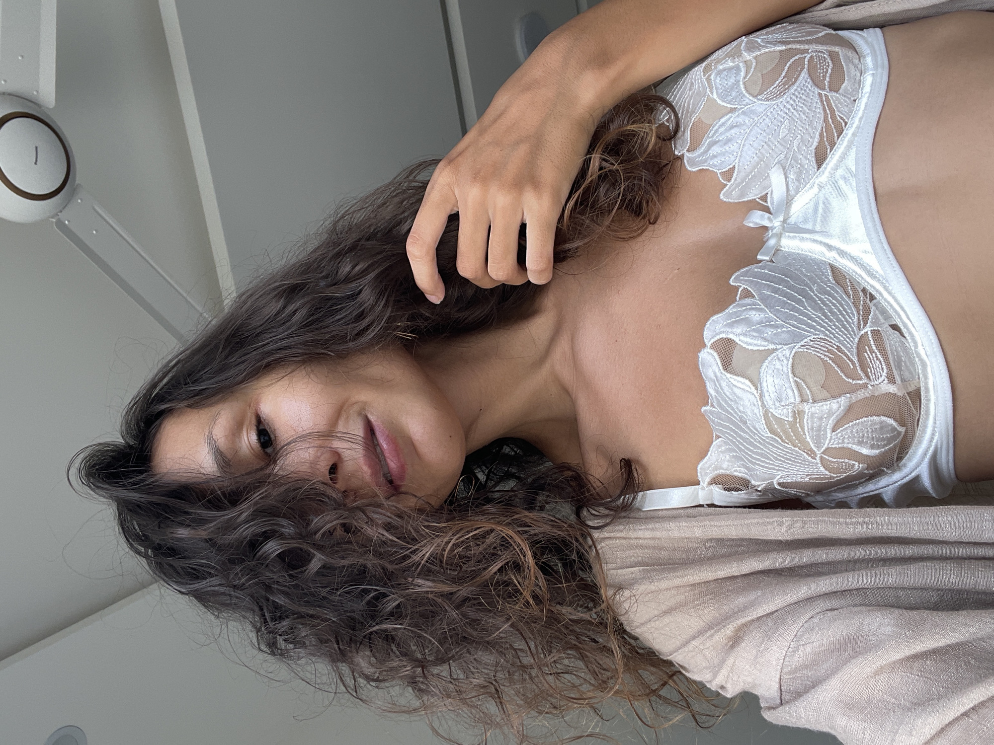 The image shows a close-up of a woman with curly brown hair, wearing a white lace bra and a beige cardigan. She is looking towards the camera and touching her hair. The background is a light-colored ceiling with a ceiling fan. The scene appears to be indoors, possibly in a bedroom or living area, and is not identifiable as being in Mũi Né, Vietnam.