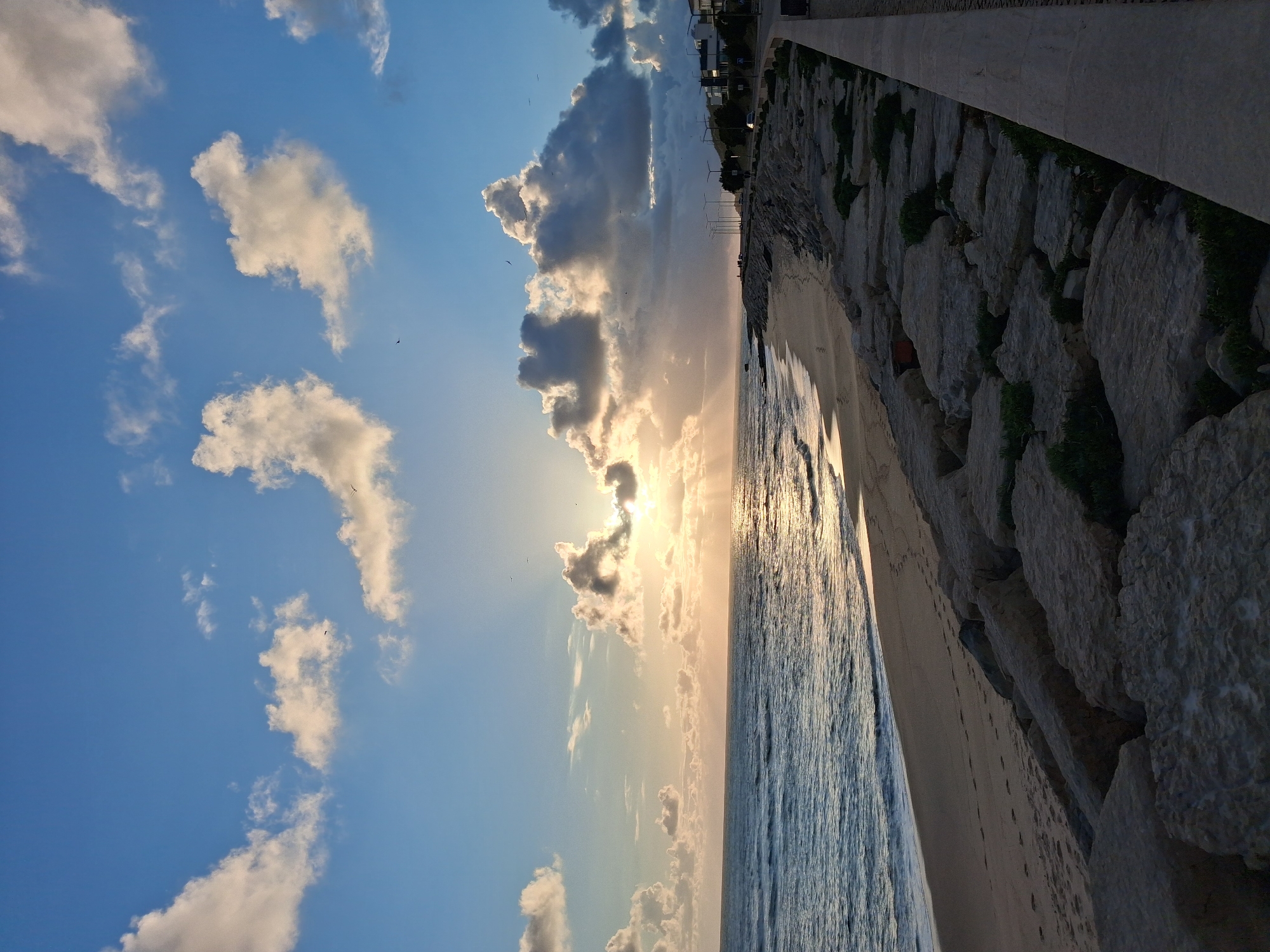 The image shows a beach scene in Figueira da Foz, Portugal. The view is rotated, presenting the sky on the left and the sea on the right. Above, a blue sky is dotted with white and gray clouds, some obscuring the sun. Birds are visible flying. The sea reflects sunlight, creating a shimmering effect. A beach runs between the sea and a stone embankment. Above the embankment, there is a sidewalk and buildings in the distance.
