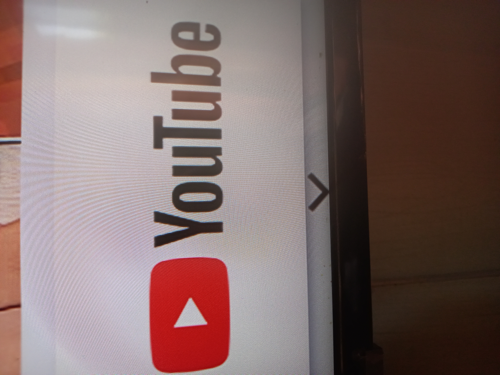 The image displays a close-up view of the YouTube logo, which consists of a red play button icon next to the word "YouTube" in dark grey lettering. The logo appears on a screen, with the pixelated texture of the display visible. Below the logo, a downward-pointing chevron icon is present, suggesting a navigation or expansion element. The screen itself is framed by a dark border at the bottom. The background above the screen appears to be a blurred image of a wooden surface. The image is taken at an angle, and the lighting suggests an indoor environment, possibly a room. There are no people or animals present in the image. The overall scene is static and focuses solely on the digital display of the YouTube logo. There is no information within the image to suggest its location in Adutiškis, Lithuania.