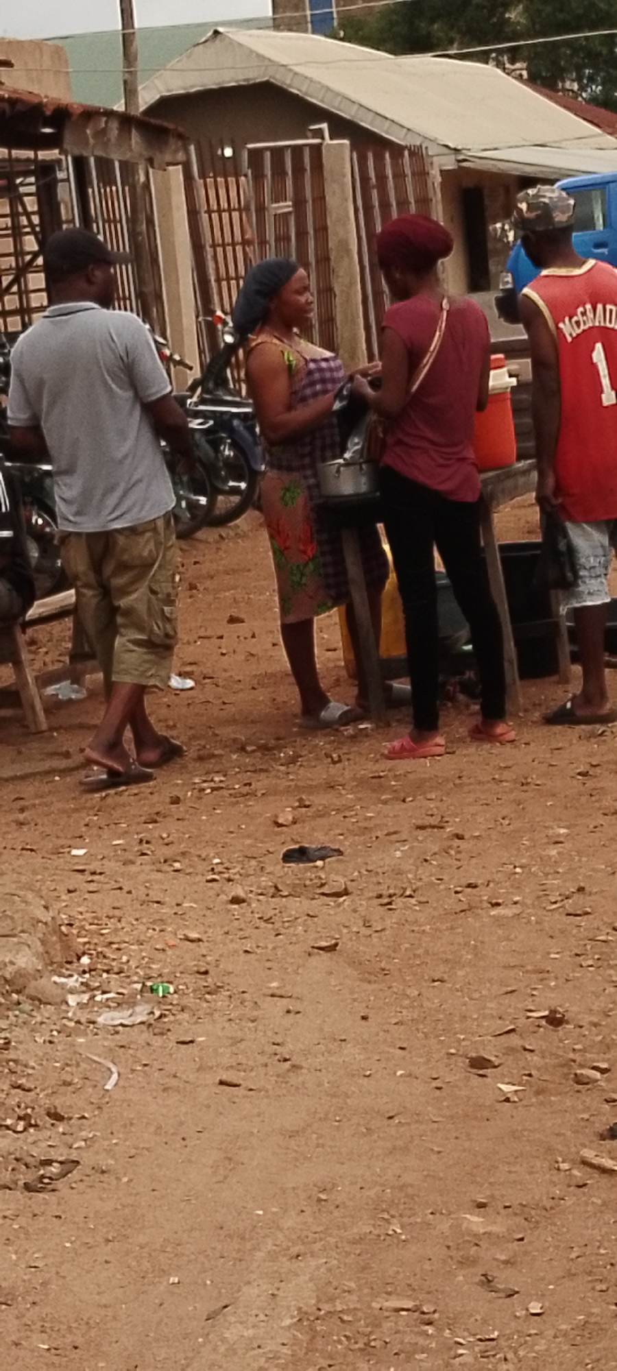 In Gboko, Nigeria, an outdoor scene captures a group of people standing on dirt ground. A man wearing a grey t-shirt and khaki shorts stands on the left, facing away from the camera. To his right, a woman in a patterned dress and headscarf is engaged in conversation with another woman dressed in a maroon top and black pants. Behind them, a man in a red basketball jersey with the number "1" and the name "McGRADY" on the back, along with camouflage-patterned headwear, stands facing left. The background reveals structures with corrugated iron roofs and barred windows, suggesting a marketplace or residential area. A blue truck is partially visible on the far right. The ground is sandy and littered with debris. Motorcycles are parked behind the individuals.