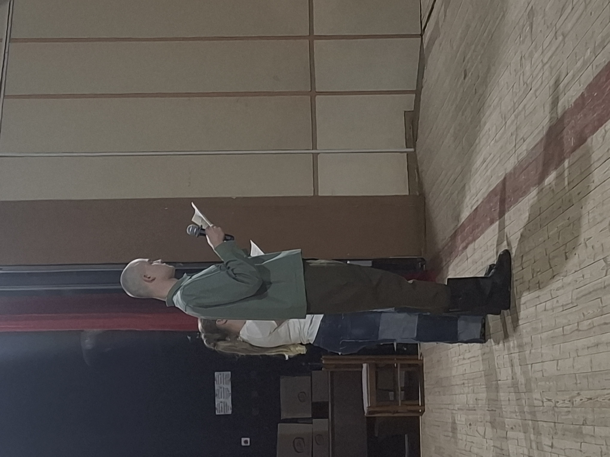 A bald man in a green jacket and brown pants stands on a stage, holding a microphone and a piece of paper. Behind him, a woman with long blonde hair wears a white shirt and blue patchwork pants. A black curtain hangs to the left of the man. Behind the woman are wooden boxes and a small wooden structure. The back and side walls are painted to resemble tile and brick. The location is inside a building, possibly in the city of Марковский, Russia.