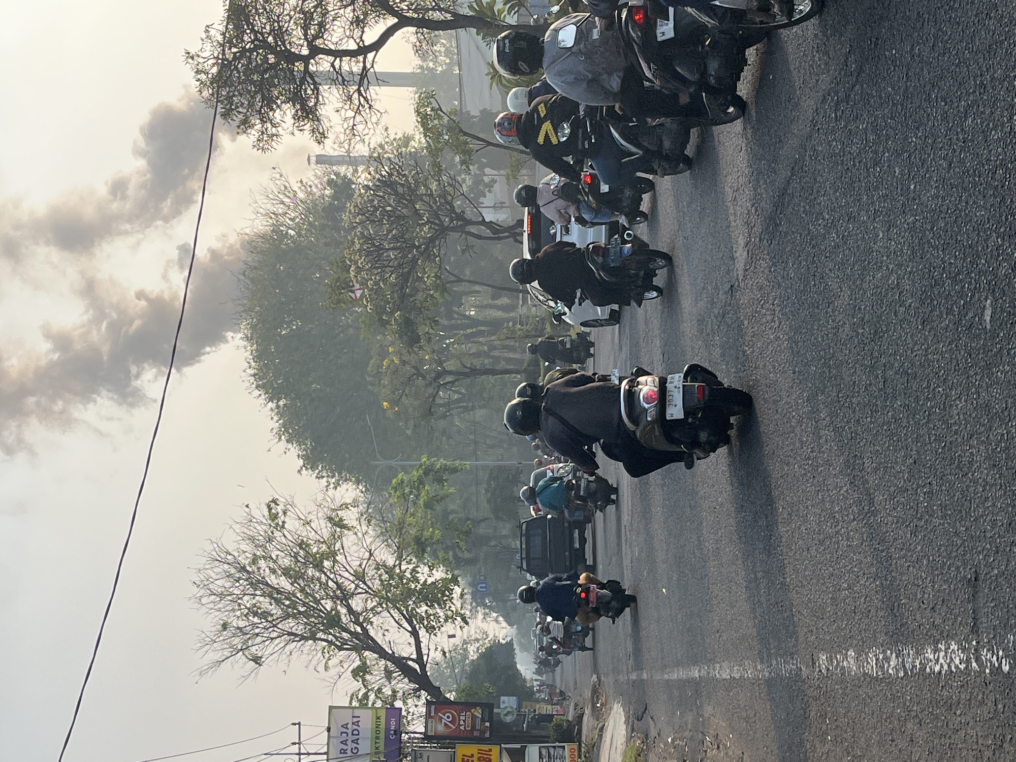 The image depicts a street scene in Candi, Indonesia, characterized by moderate traffic, predominantly motorcycles. The sky is overcast with a significant plume of dark smoke rising from industrial chimneys in the distance, suggesting air pollution. In the foreground, a road is visible with a white dashed line marking the lane. Several motorcycles with riders, all wearing helmets, are navigating the street. Some riders are looking ahead, while others are obscured from view. A black SUV is also present in the traffic. On the left side of the road, signs for "Raja Gadai" and "Candi" are partially visible, indicating commercial establishments. Trees line the sides of the road, some with sparse foliage, and buildings are present in the background, partially obscured by haze. The overall atmosphere is one of daily urban activity under a sky affected by industrial emissions.