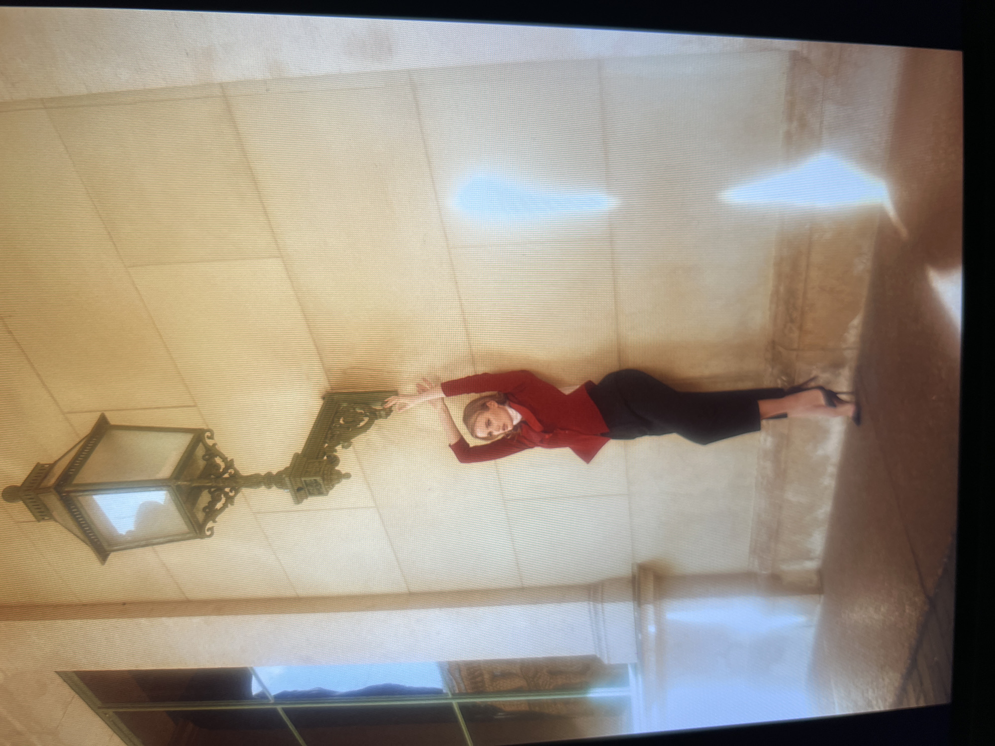 An adult female stands with her back to a stone wall, leaning against it with her arms outstretched and hands resting near a wall-mounted lamp. She is wearing a red blazer over a white top and dark trousers, with high-heeled shoes. The wall is made of large, beige stone blocks. To the left, a large window with dark frames is visible. The ground is paved with dark, rectangular stones, and there are reflections on the wet surface. A decorative, ornate lamp is attached to the wall above the woman. The lighting suggests it is daytime, with strong sunlight creating bright reflections on the wall and ground. The scene appears to be an exterior architectural setting, potentially a courtyard or facade of a building.