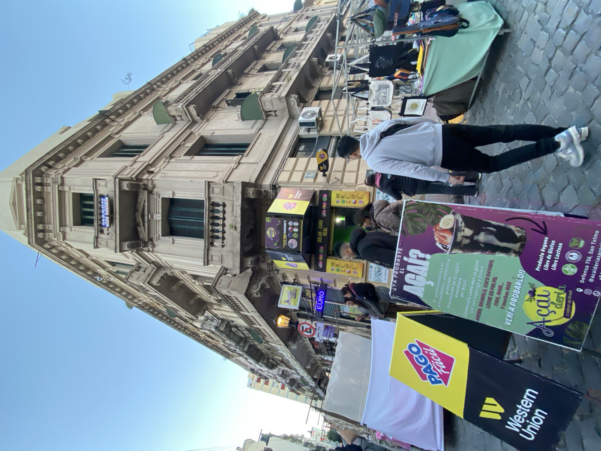 The image shows a street scene in Buenos Aires, Argentina. The building in the background is old and has a distinctive triangular shape. The street is narrow and crowded, with people walking along it. There are several vendors selling various goods, including food and souvenirs. A sign advertising "Açaí" is prominent in the foreground, along with signs for "Western Union" and "Pago Facil." Currency exchange signs, including EURO, are visible on the street.