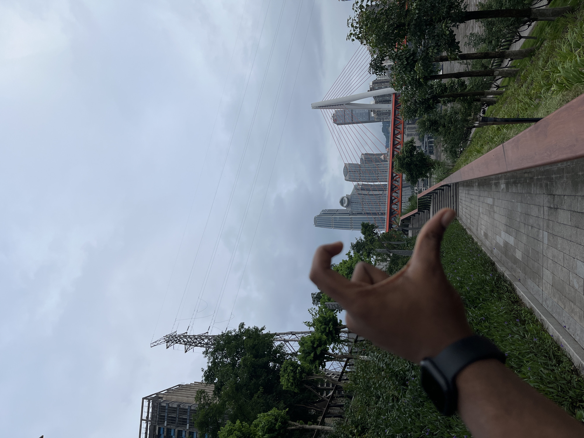 The image shows a cityscape in 江北城街道, China, under a cloudy sky. A suspension bridge with red cables stands out against the urban backdrop. High-rise buildings are visible around the bridge. In the foreground, a hand wearing a black watch is in focus, partially obscuring the scene. A paved walkway with a wooden railing is on the right side of the image, alongside greenery. Power lines and a transmission tower are also visible on the left side of the image.