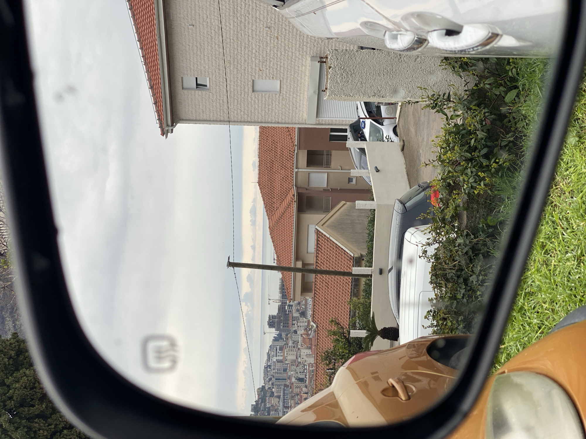 The image displays a scene in Budva, Montenegro, captured through the curved glass of a vehicle's driver-side exterior mirror, evident by the distinct heating element icon (three wavy lines) visible in the upper-left of the mirror's reflection.
In the immediate foreground, within the mirror's frame, the partial reflections of two vehicles are present: an orange-gold colored vehicle on the left, showing a door handle and a portion of its rear wheel arch, and a light-colored vehicle on the right, revealing multiple chrome door handles. Below these vehicles, wet green grass and low-lying green shrubs are visible.
The midground, reflected in the mirror, features residential architecture. A prominent light-colored, multi-story building with a textured stone facade and a red tile roof stands on the right, displaying two white-framed windows, one equipped with a closed roller shutter. Adjacent to it on the left, a lower building with a matching red tile roof is visible. Several vehicles are parked in a paved area between these structures, including a white van or car with a black roof, and a white taxi displaying a "TAXI" sign on its roof. A dark-colored vehicle is also partially visible. Green vegetation, including a small palm tree and various bushes, is present. A wooden utility pole with wires traverses the scene diagonally.
The background vista comprises a dense cityscape of Budva, characterized by numerous multi-story buildings cascading down towards the coastline. The Adriatic Sea is visible beyond the urban sprawl under an overcast grey sky. Several construction cranes are identifiable within the distant cityscape along the coast. No human figures are present in the image.