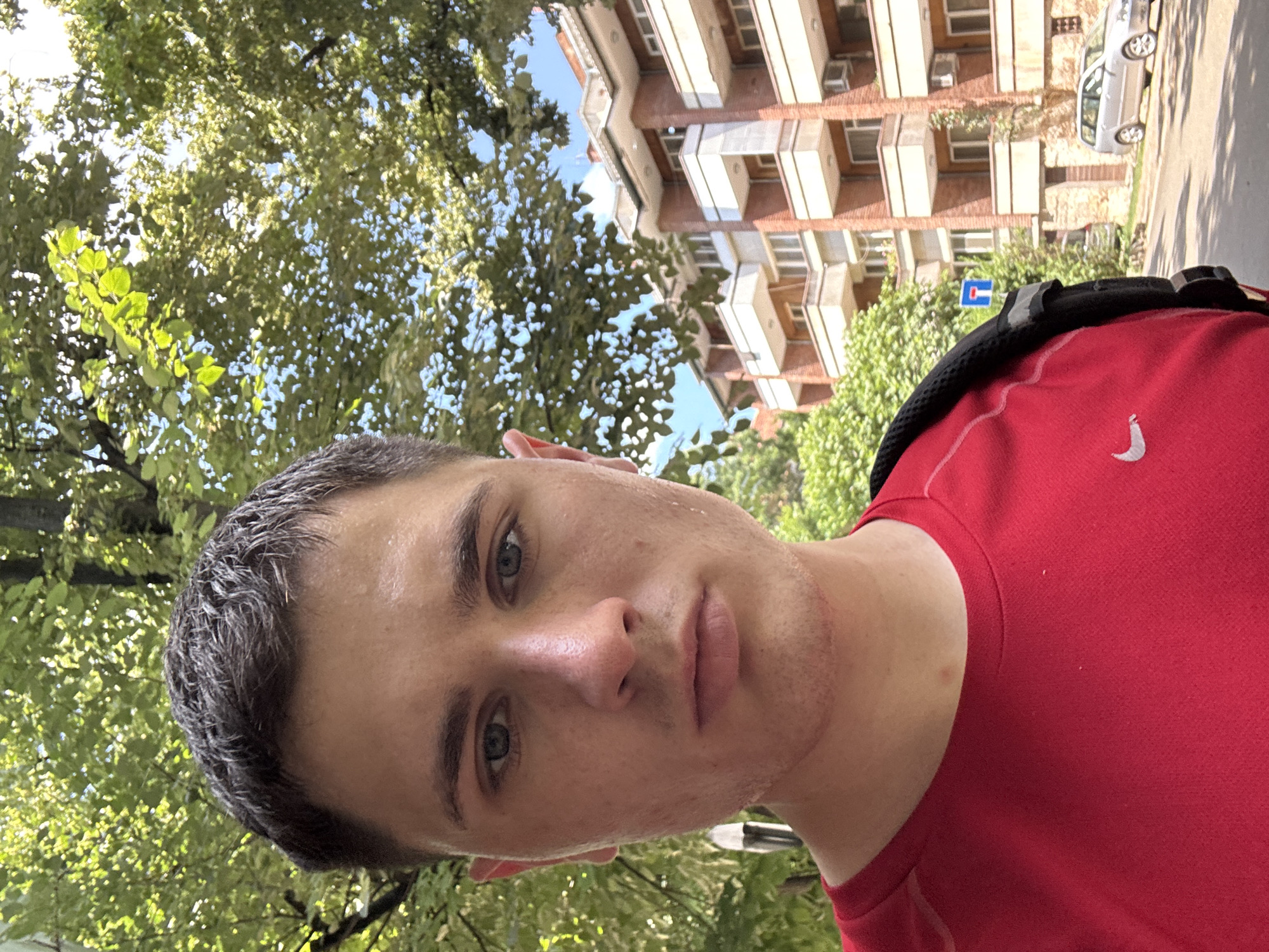 A young man with short dark hair and blue eyes is taking a selfie outdoors. He is wearing a red t-shirt and has a black backpack slung over his right shoulder. His face is slightly flushed, suggesting he may have been exercising. The background shows a tree-lined street with a modern apartment building on the right side of the frame. The building has multiple balconies and air conditioning units visible. A silver minivan is parked on the street in front of the building. A blue and white traffic sign is also visible on a pole near the building. The overall impression is a sunny day in an urban environment. This appears to be the area around G.S. Rakovski Street in Stara Zagora, Bulgaria, as indicated by the architecture and the street sign which is consistent with Bulgarian traffic signage.