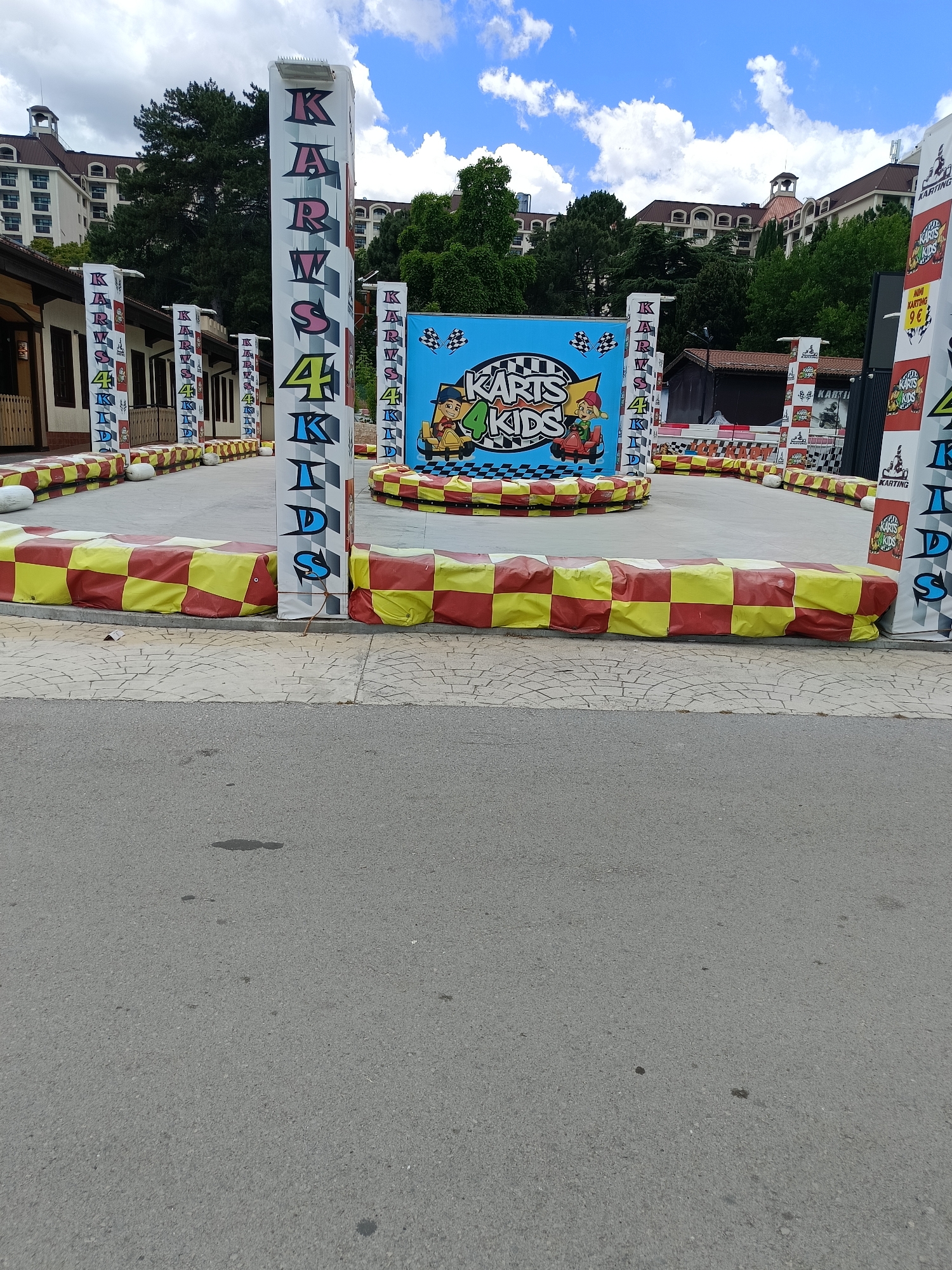 The image depicts an outdoor go-kart track for children. Several tall, white vertical banners with colorful lettering spelling "KARTS 4 KIDS" are visible along the perimeter of the track. The track itself is bordered by red and yellow checkered barriers. In the background, a large blue billboard features cartoon characters and the text "KARTS 4 KIDS". Beyond the track, there are trees and buildings, including a large multi-story hotel with arched windows and a red-tiled roof. The sky is blue with scattered white clouds. The scene is located at the Golden Sands resort in Varna, Bulgaria, a popular tourist destination known for its beaches and entertainment facilities.