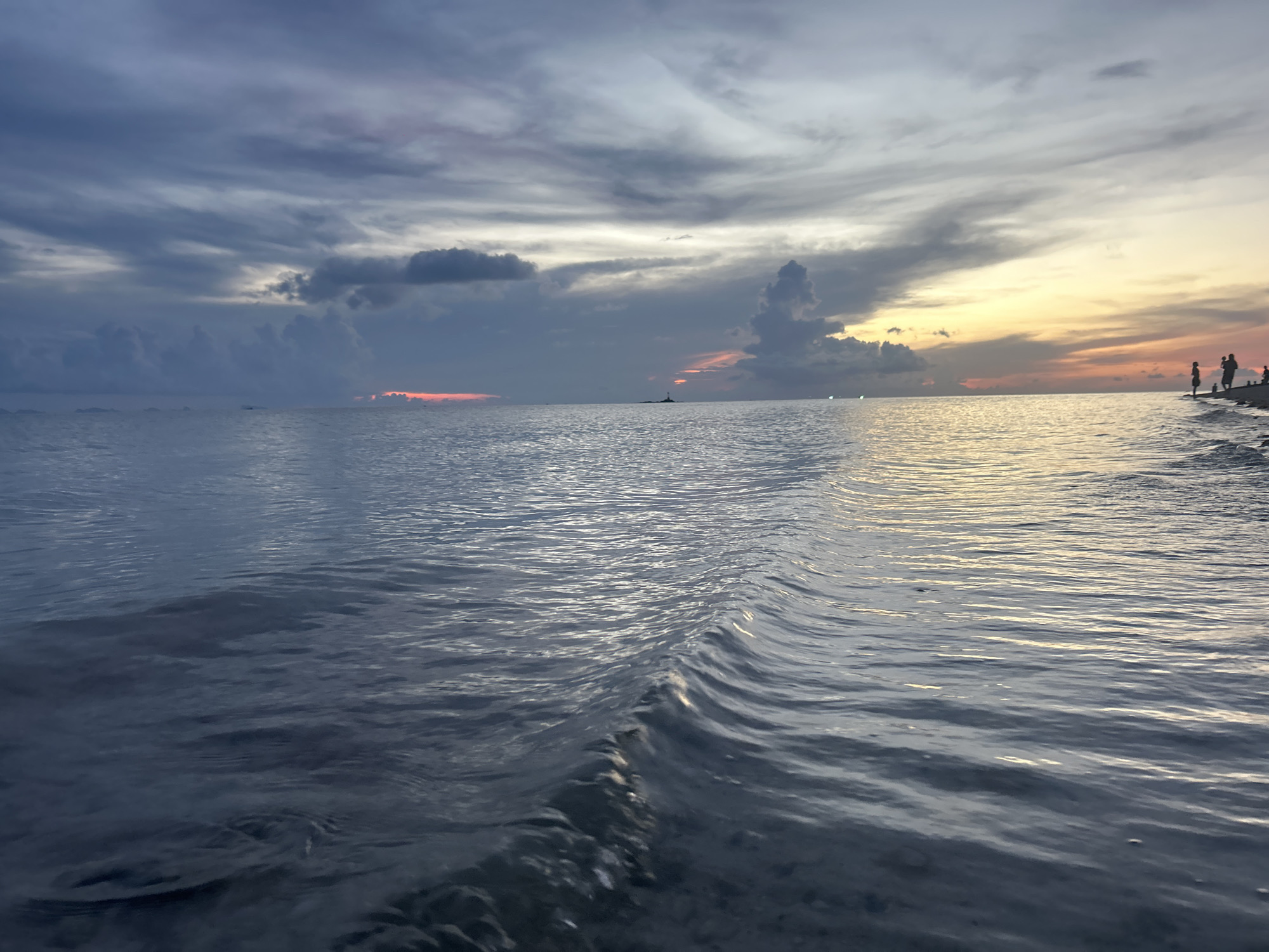 The photograph depicts a seascape at dusk in Baan Sri Thanu, Thailand. The sky is dominated by striated gray and white clouds, with faint traces of orange and yellow light visible near the horizon. The water is calm with small ripples and a few small waves. A small group of people is silhouetted on the beach in the distance on the right side of the frame.