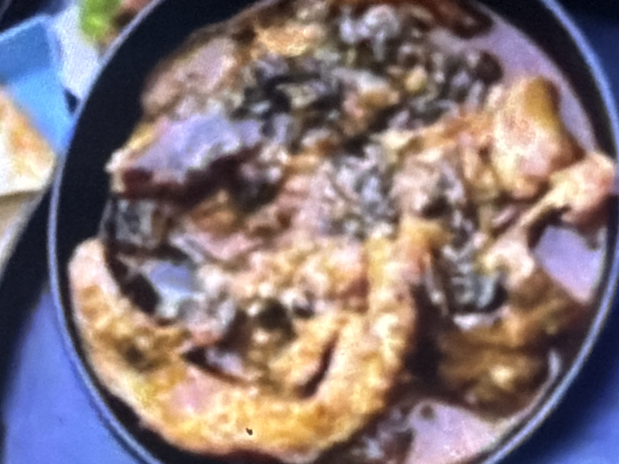 The image shows a close-up of a dish of food, possibly a stew or curry, in a black bowl. The food is a mixture of reddish-brown and yellowish-orange colors, with some darker, more textured pieces visible. The texture appears to be a sauce with chunks of meat or vegetables. The bowl is set against a blurred background with hints of blue and green, suggesting an indoor setting. There is no visible indication of the location in Bukuru, Nigeria.