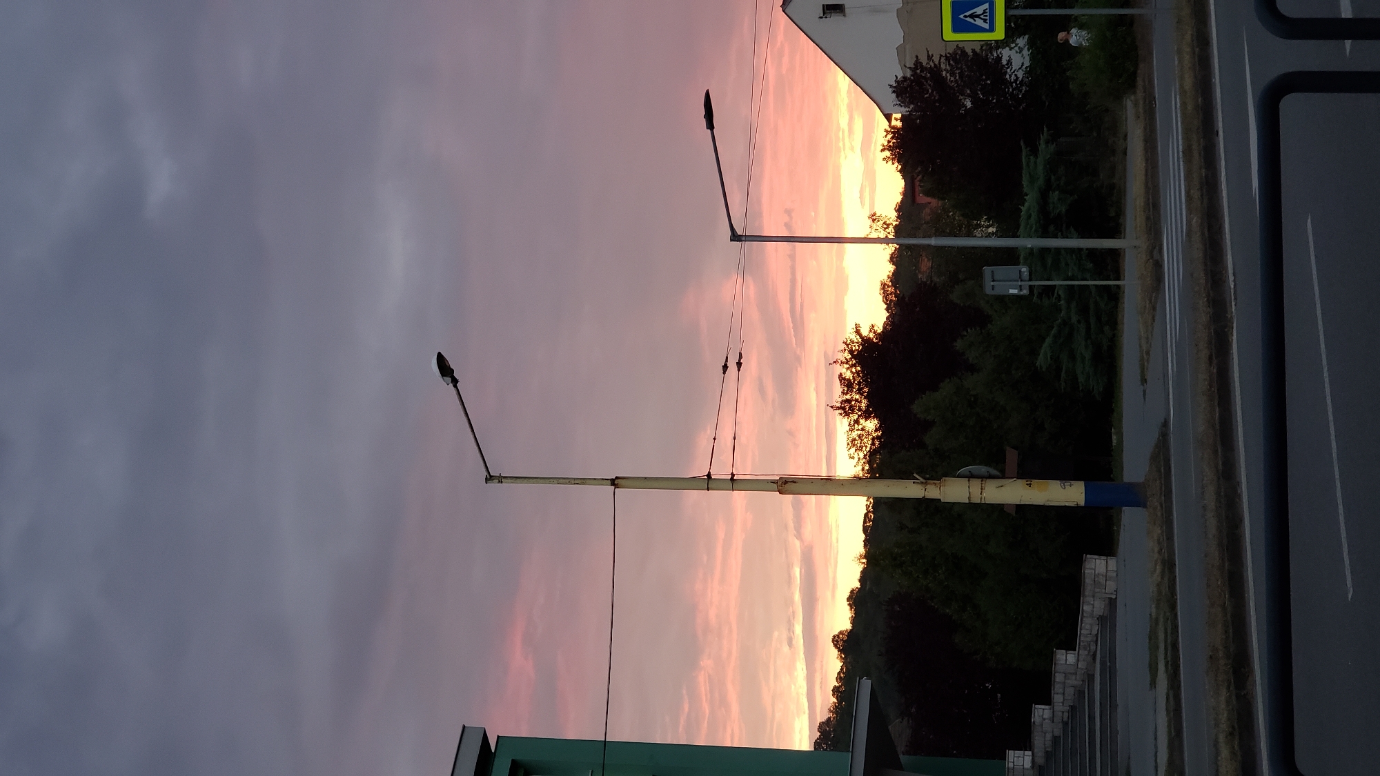 The image depicts a street scene at dusk in Prešov, Slovakia, with a vibrant sunset painting the sky in hues of pink, orange, and purple. In the foreground, a black metal railing runs horizontally across the bottom of the frame, obscuring a portion of the asphalt road marked with white lines. To the left, a section of a modern building, painted green and white, is visible with a set of concrete stairs leading up to it.
Further into the scene, two streetlights stand tall. The one on the left is a weathered yellow pole with blue accents at its base, supporting a single lamp. It is positioned on the side of the road, near a line of dark green trees. A second, more modern streetlight with a grey pole and a sleeker lamp is situated further to the right, also alongside the road.
Behind the trees, a gentle slope of a hill is visible, partially covered in dense foliage, suggesting a slightly rural or park-like setting within the city. On the right side of the image, a white building with a triangular roof is partially visible. Adjacent to this building, a yellow pedestrian crossing sign with a blue pictogram of a person walking is prominently displayed. A lone individual, casually dressed, is standing near this sign, observing the surroundings. The overall atmosphere is calm and serene, indicative of the end of the day.
