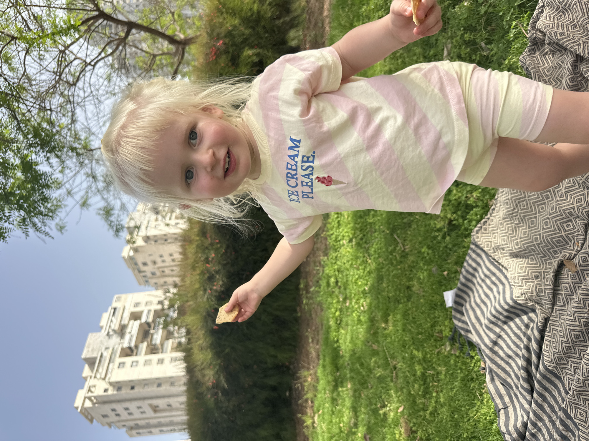 A fair-skinned, blonde-haired toddler stands on green grass. The child wears a light yellow and pink striped short-sleeved shirt with the phrase "Ice Cream Please" printed on the front, along with matching shorts. The child is holding a small, rectangular cracker in one hand and appears to be looking directly at the viewer. Behind the child, there is a large green hedge and a blue sky. In the background, multi-story white buildings are visible. A patterned blanket is partially visible on the ground to the right of the child. This image was taken outdoors in Petah Tikva, Israel.