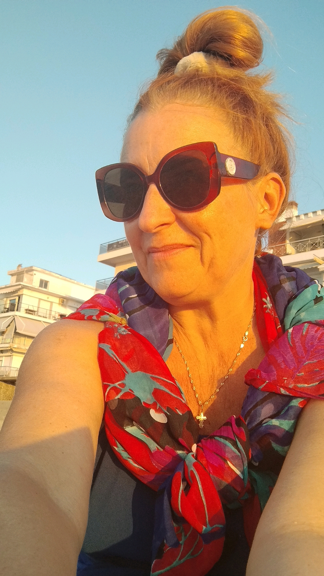 A woman with auburn hair pulled into a top knot and wearing large, dark-rimmed sunglasses, is taking a selfie outdoors. She is wearing a colorful, patterned scarf wrapped around her neck and shoulders, over a dark blue top. A thin gold chain with a small cross pendant is visible around her neck. The background shows a clear blue sky and buildings with balconies, suggesting an urban or coastal environment. The warm, golden light indicates it is likely sunset. The photo is taken at a low angle, looking up at the woman.