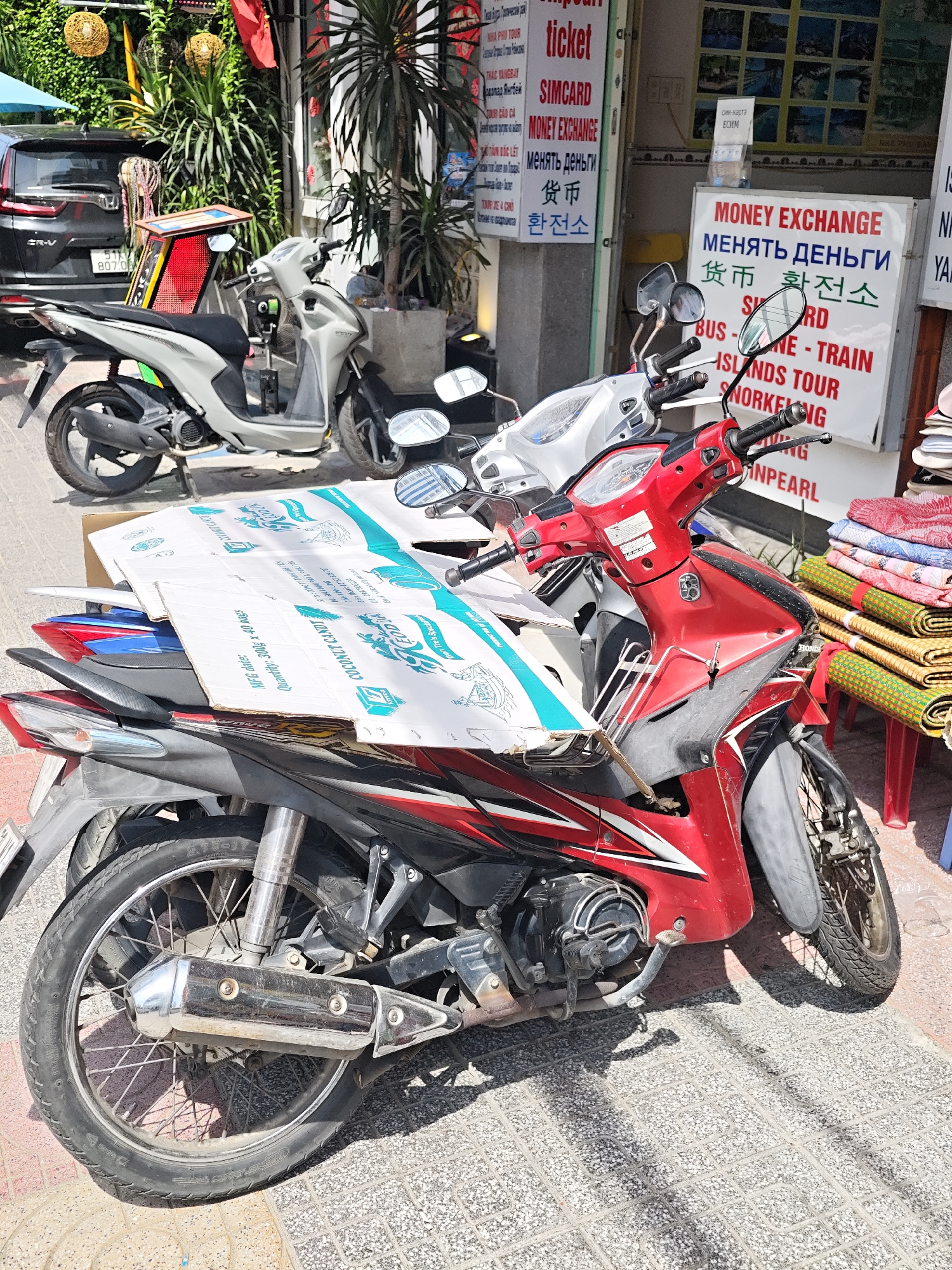 In Nha Trang Ward, Vietnam, a street scene unfolds with two scooters parked on a tiled sidewalk in front of a shop. The scooter in the foreground is red and white, with a large cardboard box resting on its seat and handlebars. This box appears to be a product box for "COCONUT CANDY". Behind it, a light gray scooter is also parked. The shop's facade features signs advertising "MONEY EXCHANGE," "SIMCARD," and various tours and services in multiple languages including English, Russian, and Korean. The background includes lush greenery and a dark colored SUV parked further down the street. The lighting suggests it is daytime.