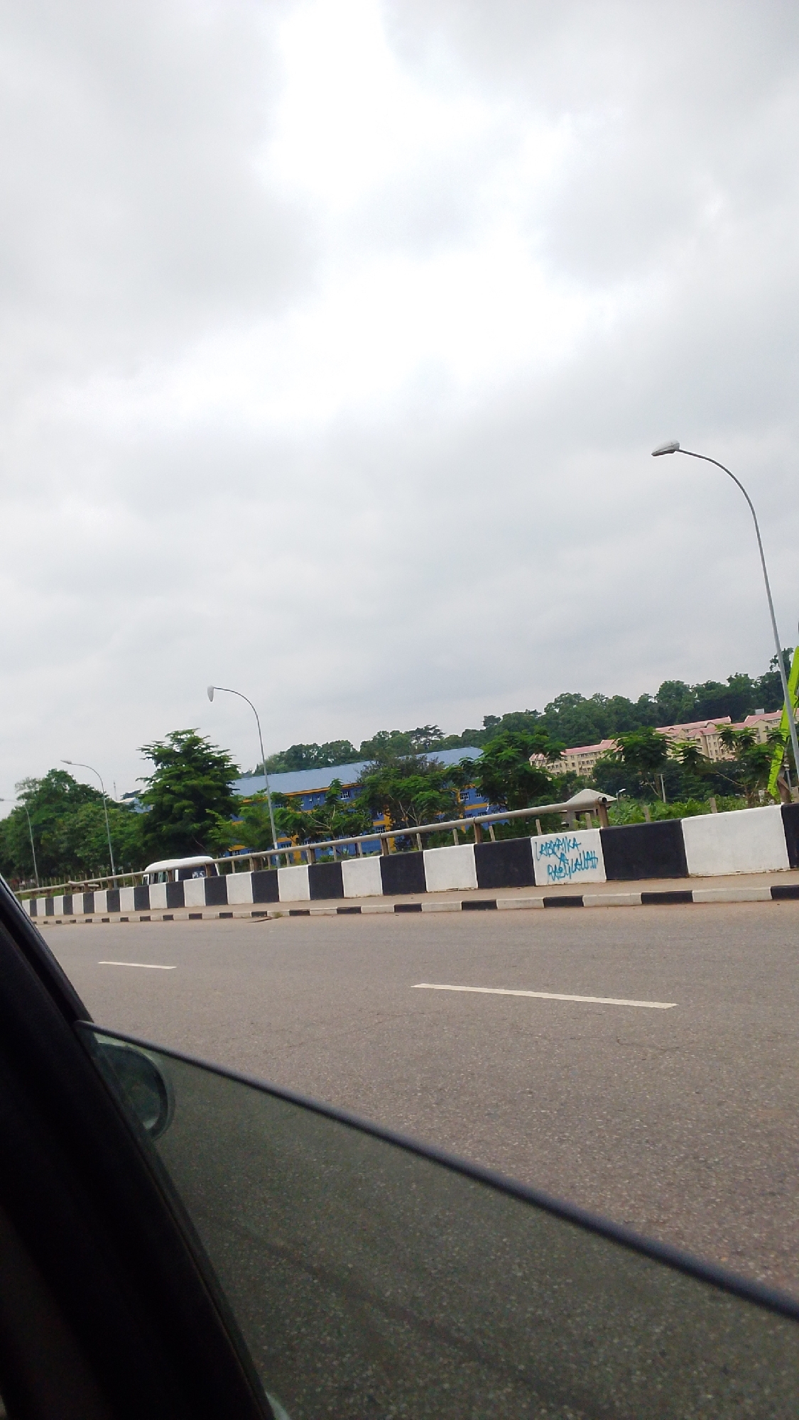An overcast day in Aso, Nigeria, shows a highway with a black and white striped barrier. Behind the barrier, a white bus is visible amongst trees and buildings. A street light is visible on the right side of the image. The foreground shows the interior of a car with the window partially down.