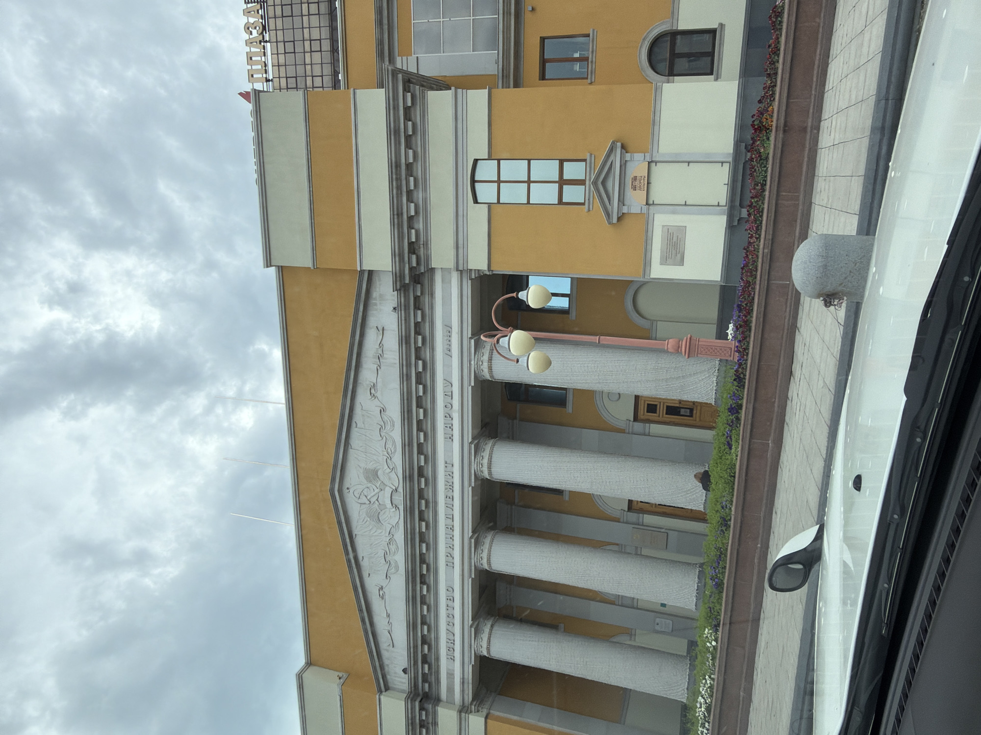 The image shows the exterior of a building with a grand facade. The building is painted in yellow and cream colors, with large white columns and an ornate pediment above them. Inscriptions are visible on the facade, in Russian text. A lamppost with two globes stands in front of the columns. The building appears to be located on a slightly sloping street, with a flowerbed in front of it. A car's interior, including the dashboard and a side mirror, is visible in the foreground, suggesting the photo was taken from inside a vehicle. A sign with "ПЛАЗА" (Plaza) is visible on the upper right part of the building. This building is the Khabarovsk Regional Palace of Culture trade union, located in Khabarovsk, Russia.