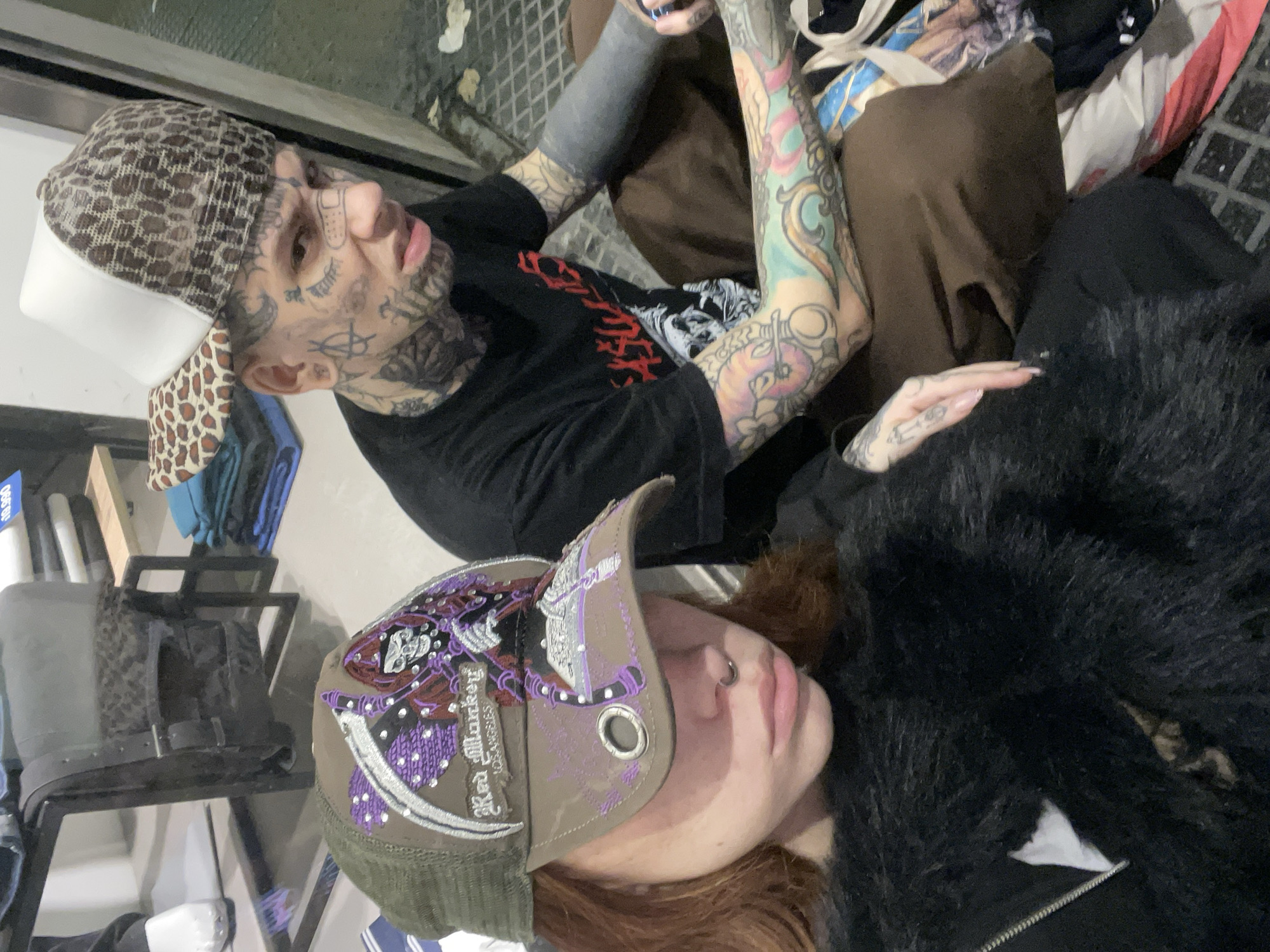 Two individuals are present in the scene. One individual, with extensive face and arm tattoos and wearing a leopard-print baseball cap and a black T-shirt, is sitting with their legs crossed. The other, who is wearing a baseball cap with purple and silver embellishments and a black furry jacket, is sitting beside them. The background includes shelving with items such as clothes and accessories, as well as what appears to be a metal grate or flooring. There is no specific information in the photo that indicates the location is Buenos Aires, Argentina.