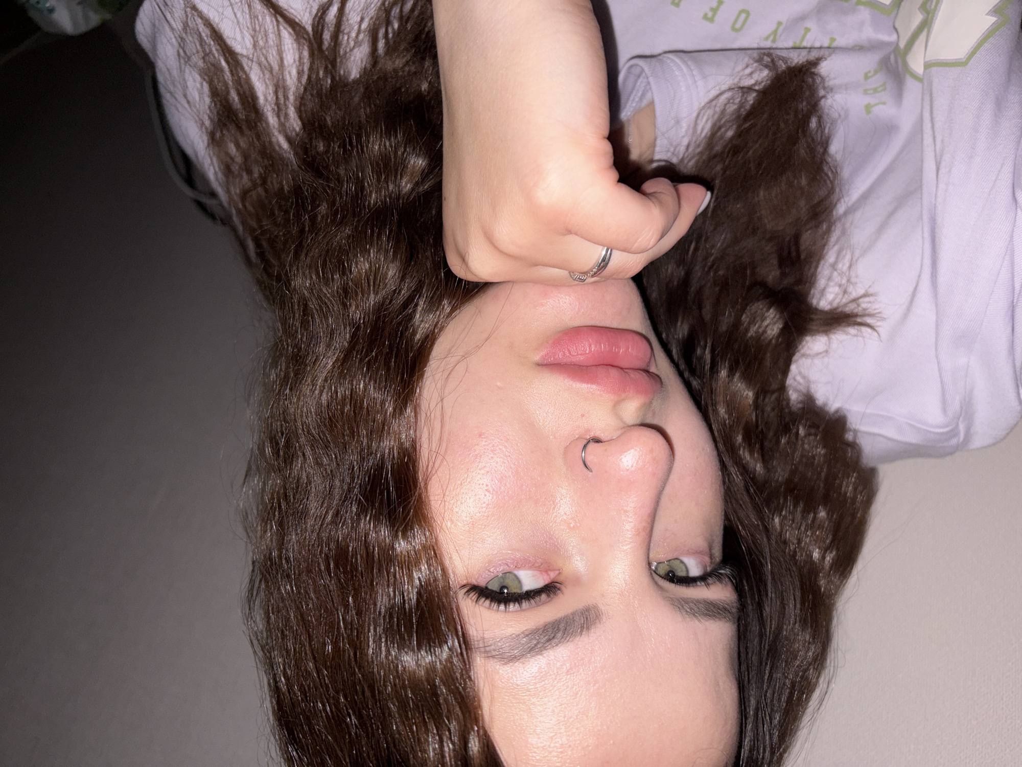 A young woman with dark brown, curly hair is lying down, facing the camera. Her right hand is raised and positioned near her nose. She has light skin, green eyes, and a nose ring. She is wearing a light purple t-shirt. The background is a solid, neutral color. There is no indication of a specific location, including Krasnodar, Russia.