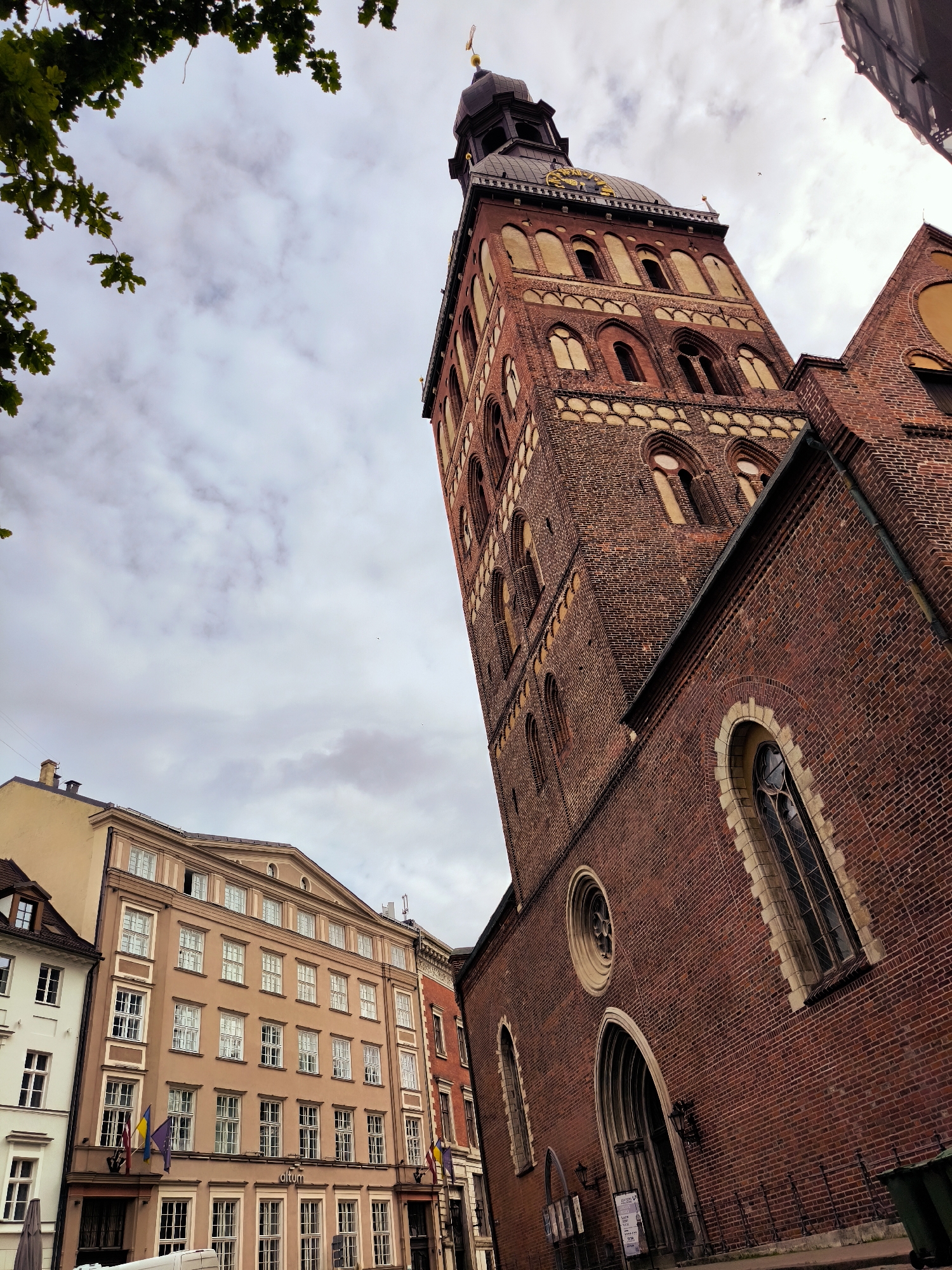 The image shows a low-angle view of the Riga Cathedral's brick tower and adjacent buildings in Riga, Latvia. The sky is overcast with scattered clouds. Green tree branches frame the upper left corner of the image. The cathedral tower is tall and constructed of dark red brick, featuring multiple levels with arched windows and decorative brickwork. A clock face is visible near the top of the tower. To the left of the tower is a multi-story building with a light brown facade, numerous white-framed windows, and a triangular pediment at the top. Several flags are displayed on this building. Further to the left, a white building with dormer windows is partially visible. The foreground shows a sidewalk and part of a street with some trash bins. The scene depicts an urban environment with historical architecture.