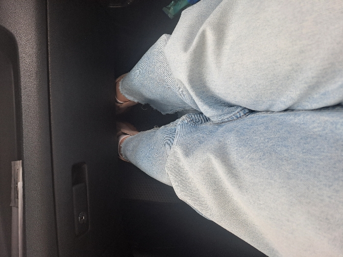 The image displays a first-person perspective captured from within a vehicle interior, looking downward at the occupant's lower body and forward into the dashboard area. The occupant's legs are clothed in light-wash blue denim jeans, which feature frayed or distressed hems above the ankles. The feet are encased in light-colored, open-toe shoes or sandals. Directly ahead, the vehicle's dark-colored dashboard or lower glove compartment panel is visible, characterized by a textured surface. A dark glove compartment latch, incorporating a silver-toned keyhole or USB port, is centrally located on this panel. Above the glove compartment, a narrow horizontal slot contains a white, rectangular item. The floor area beneath the feet appears dark. A partially obscured green or blue object is visible on the far right side of the frame. The specific location in the city of Obukhovo, Russia, is not ascertainable from the visual evidence within the image.
