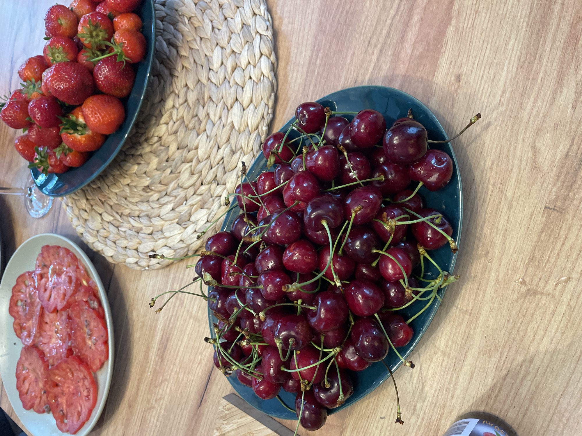 A high-angle, medium shot shows a wooden table with three plates of fruit, a woven placemat, and a glass. In the foreground, a dark green oval plate is filled with a large pile of fresh cherries. The cherries are dark red with green stems. To the left and slightly behind the cherries, a light-colored plate contains several slices of red tomato, seasoned with what appears to be salt and pepper. To the right and further back, another dark green plate is overflowing with bright red strawberries, some of which have green leaves attached. A woven placemat, made of light-colored straw, sits beneath the strawberry plate. A partial view of a stemmed glass is visible in the upper left corner of the frame. The table surface is made of light-brown wood planks.