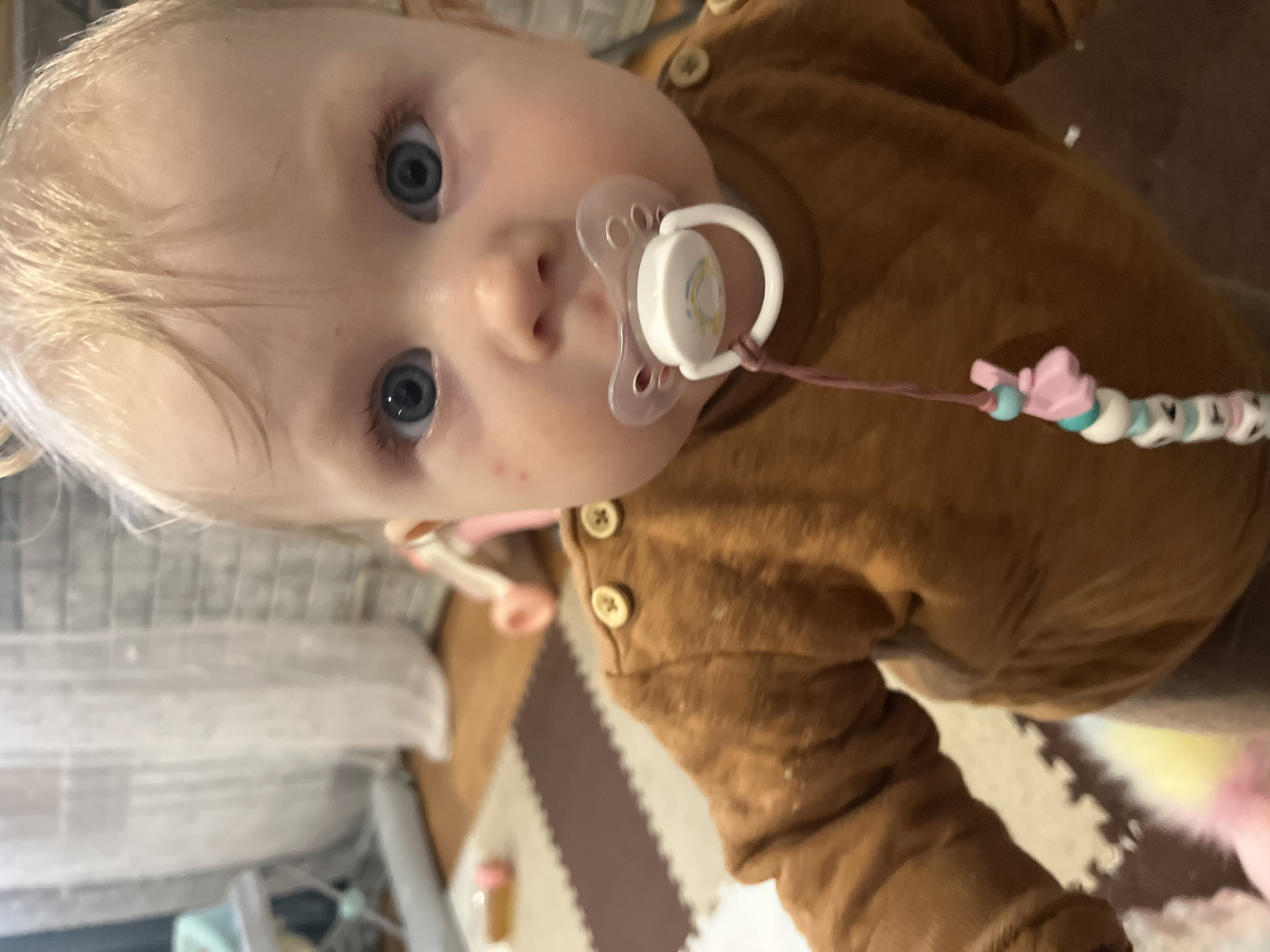 A close-up, indoor photograph features a young baby, estimated to be between six months and one year old, looking directly at the camera. The baby has fair skin, light blonde hair, and striking blue eyes that are wide and attentive. A clear plastic pacifier is in the baby's mouth, attached to a pacifier clip adorned with colorful beads, including some white square beads with visible dark letters, such as "KTA" or similar. The baby is dressed in a long-sleeved, rust-brown top featuring light-colored buttons on the shoulder. Small reddish spots are visible on the baby's left cheek.
The background, mostly out of focus, reveals an indoor setting with interlocking foam play mats on the floor, suggesting a home environment or dedicated play area. The lighting appears soft, consistent with an indoor daytime or evening setting. The baby's direct gaze conveys curiosity and engagement. This scene takes place in Vesyoloye, Russia.