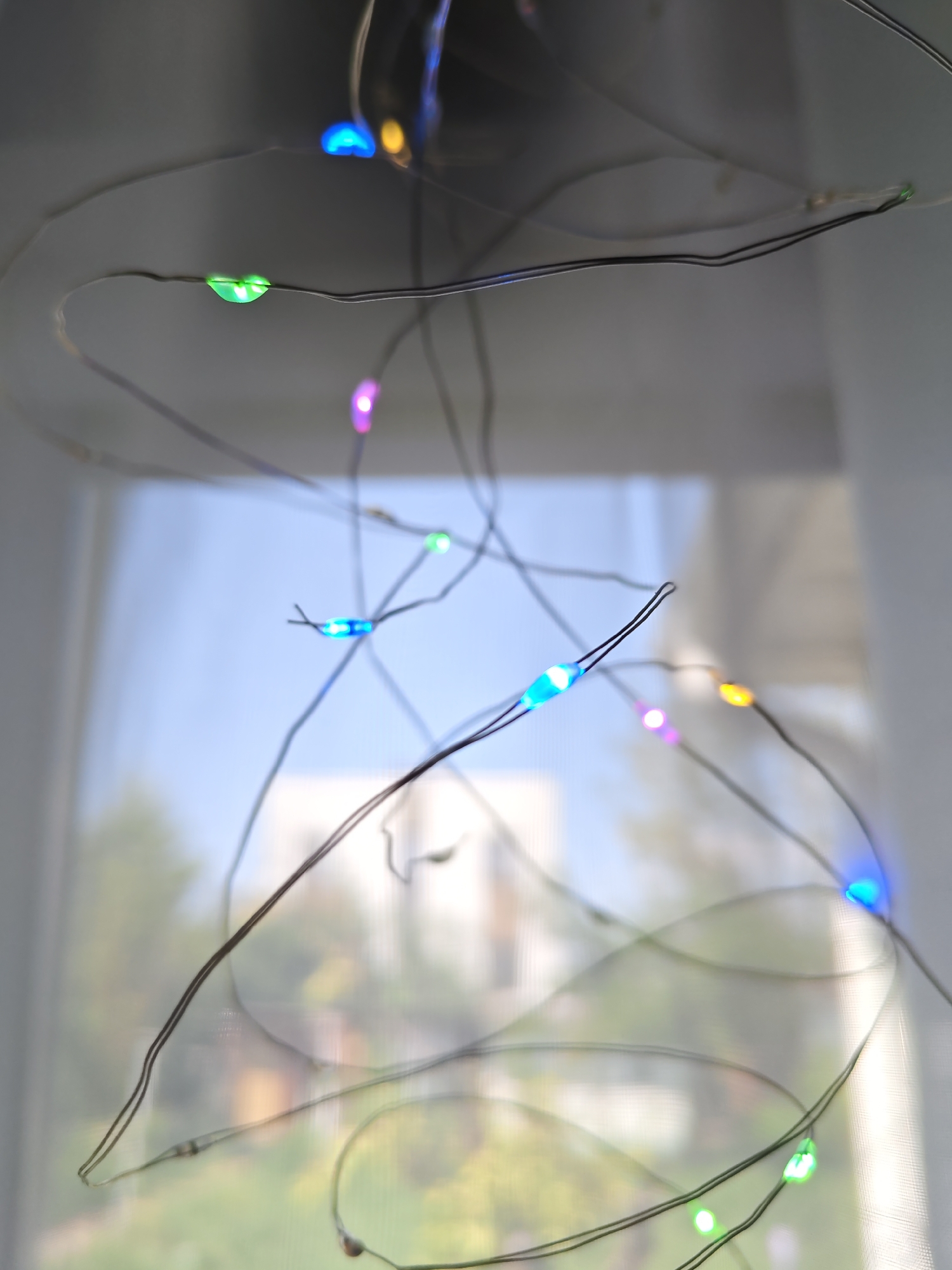 The image shows a close-up of a string of colorful LED lights against a blurred background. The lights are small, bulb-shaped, and illuminated in various colors including blue, green, purple, and yellow. They are attached to a thin, dark wire that is wound in a tangled manner. The background appears to be a window with sheer curtains, through which glimpses of greenery and a pale blue sky are visible, suggesting an outdoor scene beyond. There are no people or discernible landmarks indicating a specific location in Rostov-on-Don, Russia. The focus is on the abstract pattern and colors of the lights.