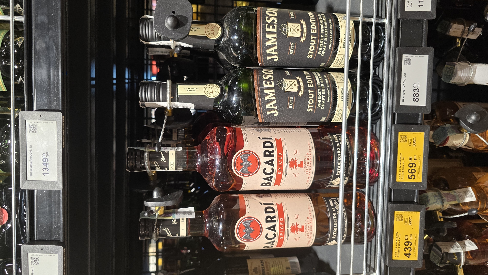 The image shows a retail shelf displaying bottles of alcohol in a store. In the foreground, there are price tags for Bacardi Spiced rum bottles. One tag indicates a price of 439.00 UAH for 0.7L and another shows 569.00 UAH for 1L. To the right, a price tag displays 883.00 UAH for 0.7L of Jameson whiskey. The bottles are arranged on a metal shelf with other alcoholic beverages. The environment appears to be a well-lit store aisle, possibly a supermarket or liquor store. The image does not provide specific details to pinpoint the location in Zhytomyr, Ukraine, but it is a typical retail display of alcoholic beverages. There are no people or interactions visible in the image.
