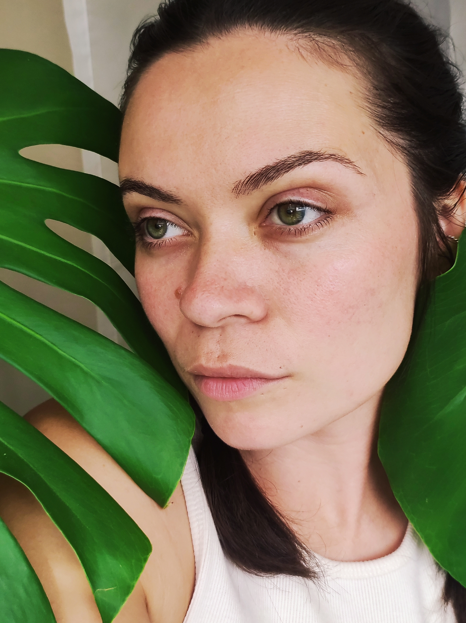 A close-up, medium shot shows a woman's face, framed by two large, bright green leaves of a monstera plant. The leaves are positioned on either side of her face, obscuring her left cheek and partially covering the right side of her head and neck. The woman has fair skin, light brown hair pulled back from her face, and striking green eyes. Her gaze is directed to her left, away from the camera, with a neutral expression. She is wearing a white ribbed tank top. The background is a soft, neutral-toned wall. There is no indication of the city of Chaikovsky, Russia, in this image.