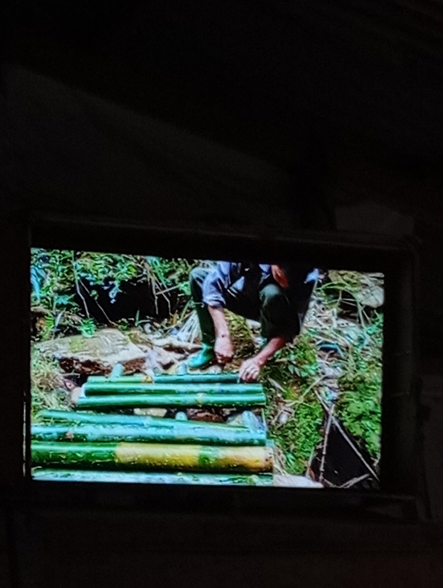 The image displays a projection on a dark surface, showing a close-up of a person, likely a man, wearing dark pants and green boots, crouching and interacting with a series of green bamboo poles arranged horizontally. The background of the projection features lush green foliage and rocks, suggesting an outdoor, natural environment. One of the bamboo poles, towards the bottom, appears to have a yellow hue. The scene is dimly lit, with the projection being the primary source of illumination. The specific location in Lhokseumawe, Indonesia cannot be determined from the visual information provided.