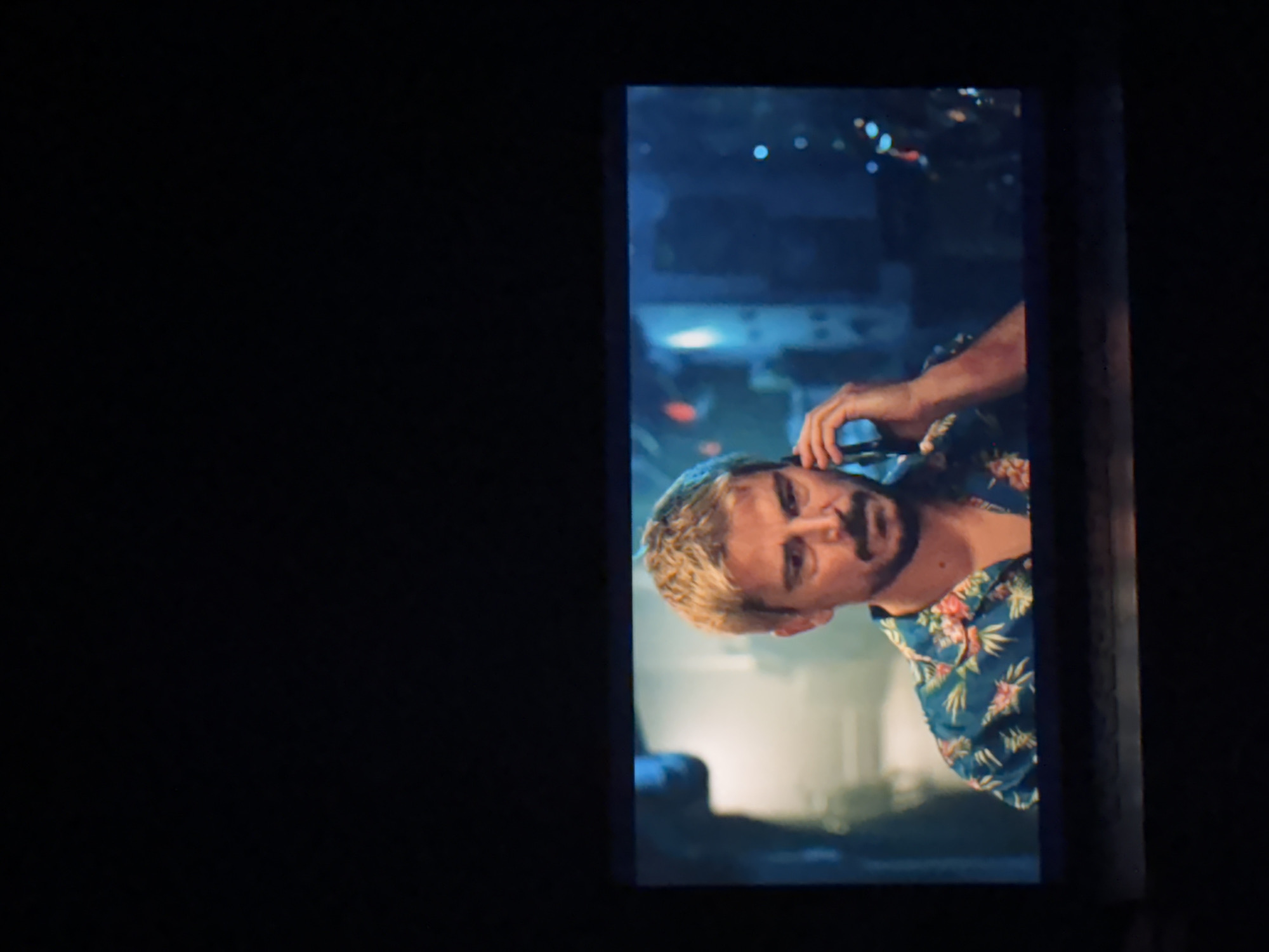 The image captures a vertically oriented screen displaying a scene, surrounded by a largely dark, unlit environment. The screen shows a man with light-colored, possibly blond, hair and a beard, wearing a blue short-sleeved shirt with a vibrant floral pattern, reminiscent of a Hawaiian shirt. He is holding a dark-colored mobile phone to his right ear with his right hand, seemingly in conversation. His expression appears serious or pensive, with his gaze directed slightly off-camera.
The background of the scene on the screen is blurred, suggesting a nighttime, possibly urban or coastal setting, consistent with Budva, Montenegro. There are bokeh effects from distant lights, indicating artificial light sources in the dark. The ambient lighting on the man's face and shirt is clear, contrasting with the darker, slightly bluish background. The overall impression is a candid shot of someone on a phone call at night. There is no visible text in the image.