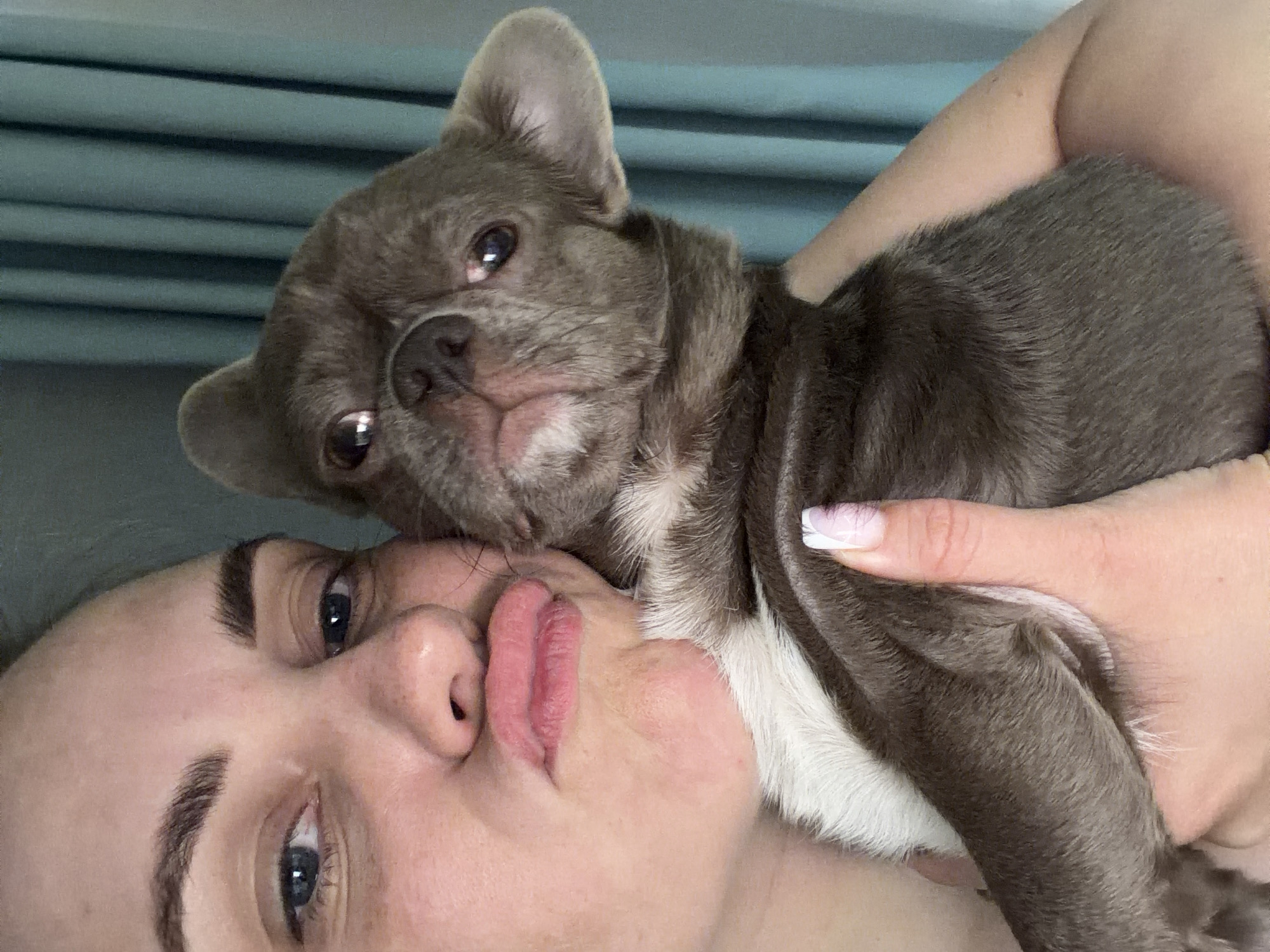 A woman lies on her back, holding a gray French bulldog puppy. The puppy is resting its head on her face. The woman looks up towards the camera with blue eyes and plump lips. She has well-defined eyebrows. Her right hand is visible, with a manicured nail painted with a white tip. Behind the pair is a teal-colored, textured background. There is no indication of the location being Votkinsk, Russia.