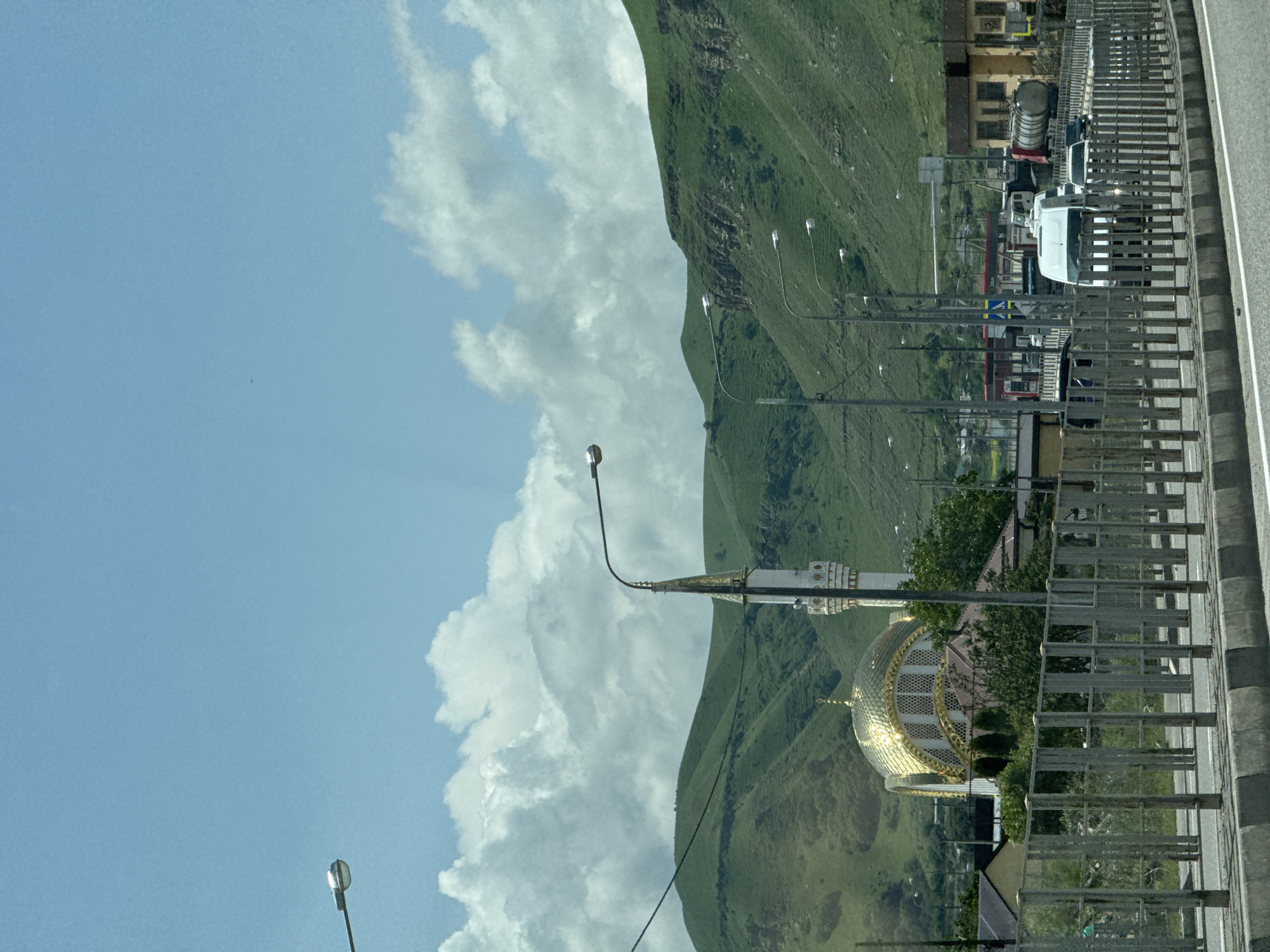 A mosque with a golden dome and a white minaret stands prominently against a backdrop of rolling green hills under a bright blue sky with scattered white clouds. The mosque is located in Krasnogorskaya, Russia. In the foreground, a metal fence runs along a road where several vehicles are visible, including a white van, a truck with a tanker trailer, and other cars. The road is bordered by a concrete barrier. Streetlights are positioned along the road. The overall scene depicts a landscape with architectural elements and traffic, set against a natural mountainous environment.