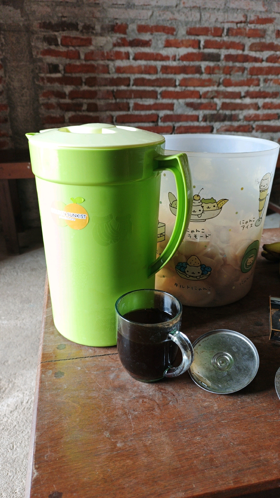 A clear glass mug filled with a dark liquid, likely coffee, sits on a wooden table. Next to the mug is a silver lid, possibly for the mug or another container. Behind the mug and lid is a large, lime green plastic pitcher with a white lid and a green handle. To the right of the green pitcher is a translucent white plastic container decorated with cartoon cat illustrations. The background consists of a brick wall. The scene is likely located in Ngranti, Indonesia, as indicated by the presence of objects commonly found in Indonesian households and the mention of the location.