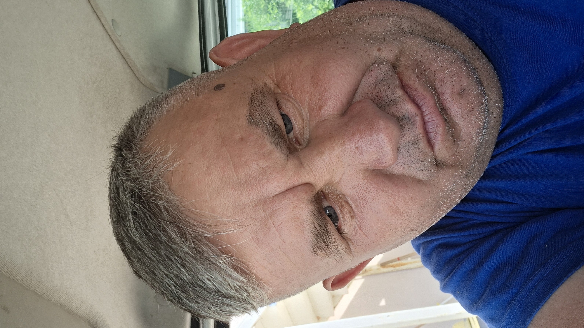 This media file is a close-up, horizontally oriented photograph of a middle-aged man's face. He has graying hair, stubble on his face, and fair skin with some visible pores and wrinkles. His eyes are blue and appear to be looking slightly off-camera. A mole is visible on his left cheek. He is wearing a blue t-shirt. The background includes a light-colored textured surface on the left, possibly an interior car headliner, and a blurred green outdoor scene visible through what appears to be a window on the upper right. The man's head is tilted, and the lighting suggests it is daytime. There is no visible text in the image. The overall impression is a casual, candid portrait.