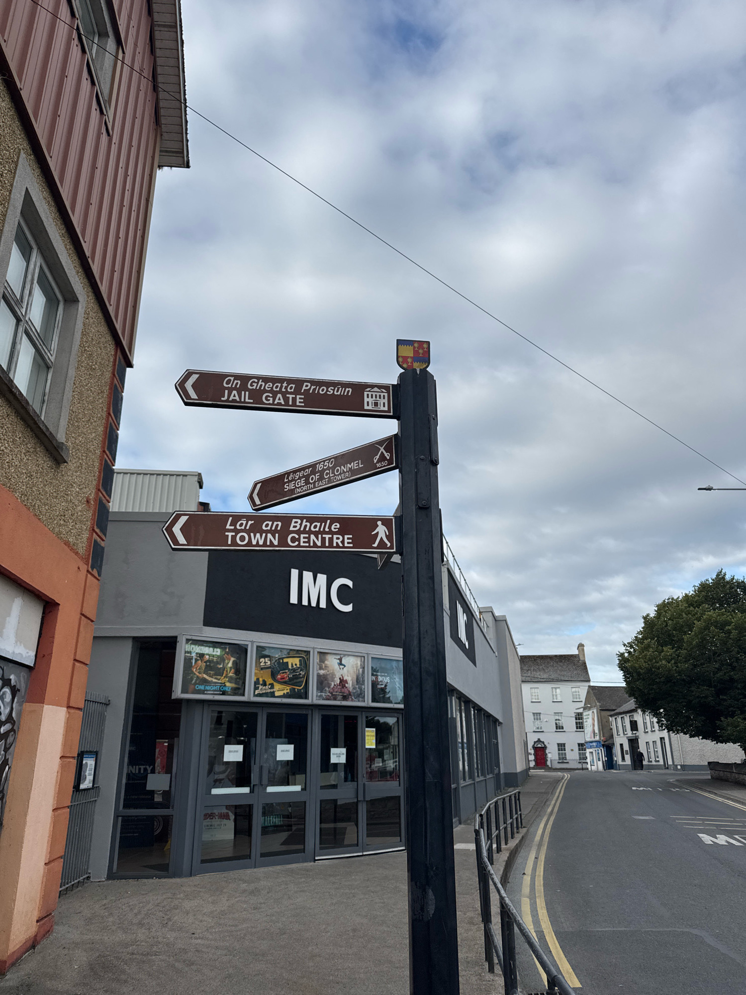 The image shows a street scene in Clonmel, Ireland. A signpost with multiple brown directional signs points in different directions. The top sign reads "On Gheata Priosuin JAIL GATE". Below it, another sign reads "Léigear 1650 SIEGE OF CLONMEL (NORTH EAST TOWER)". The third sign says "Lár an Bhaile TOWN CENTRE" with a pictogram of a person walking. The signpost is topped with a shield bearing a coat of arms. To the left of the signpost is a building with a corrugated iron facade, and further to the left, part of a building with orange and gray textured walls. In front of the signpost is a modern building with large glass doors and windows, displaying movie posters and the "IMC" logo. The street is paved and has yellow lines marking lanes. In the background, there are more buildings, including a white building with a red door and a large green tree on the right side of the street. The sky is cloudy.