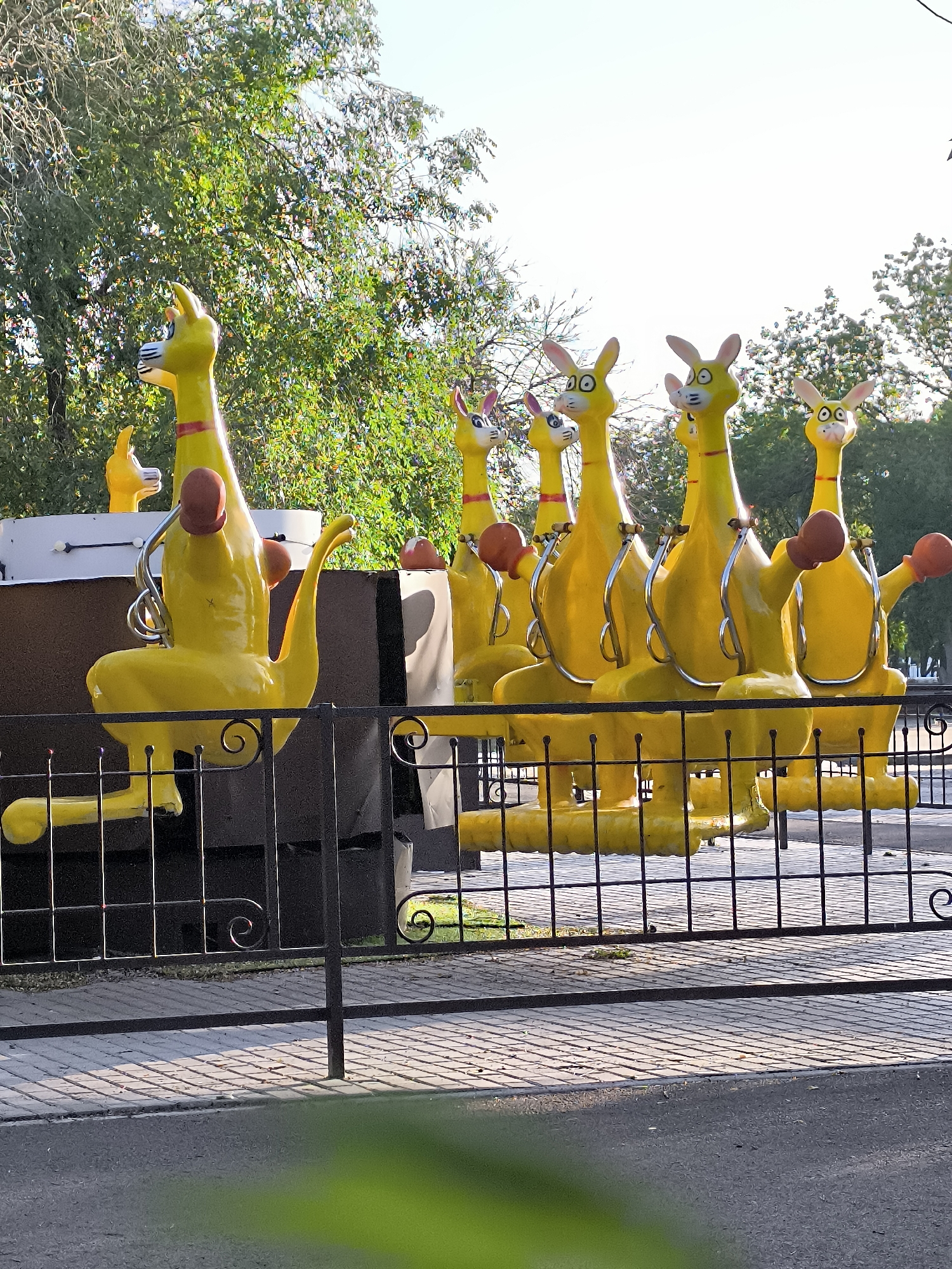 A row of vibrant yellow kangaroo figures, designed as amusement park rides, are lined up outdoors behind a black metal fence. The kangaroo figures are all in a seated position with their arms bent, as if in mid-motion. Each kangaroo has large eyes and long ears. Some figures are equipped with maroon boxing gloves on their hands. The background consists of lush green trees and a clear sky, suggesting a sunny day. The ground in front of the fence is paved with grey interlocking stones, and a portion of an asphalt road is visible in the foreground, out of focus. The scene appears to be a section of an amusement park or a similar recreational area within the city of Qarağandı, Kazakhstan, specifically near the city administration building. No people are visible in the frame.