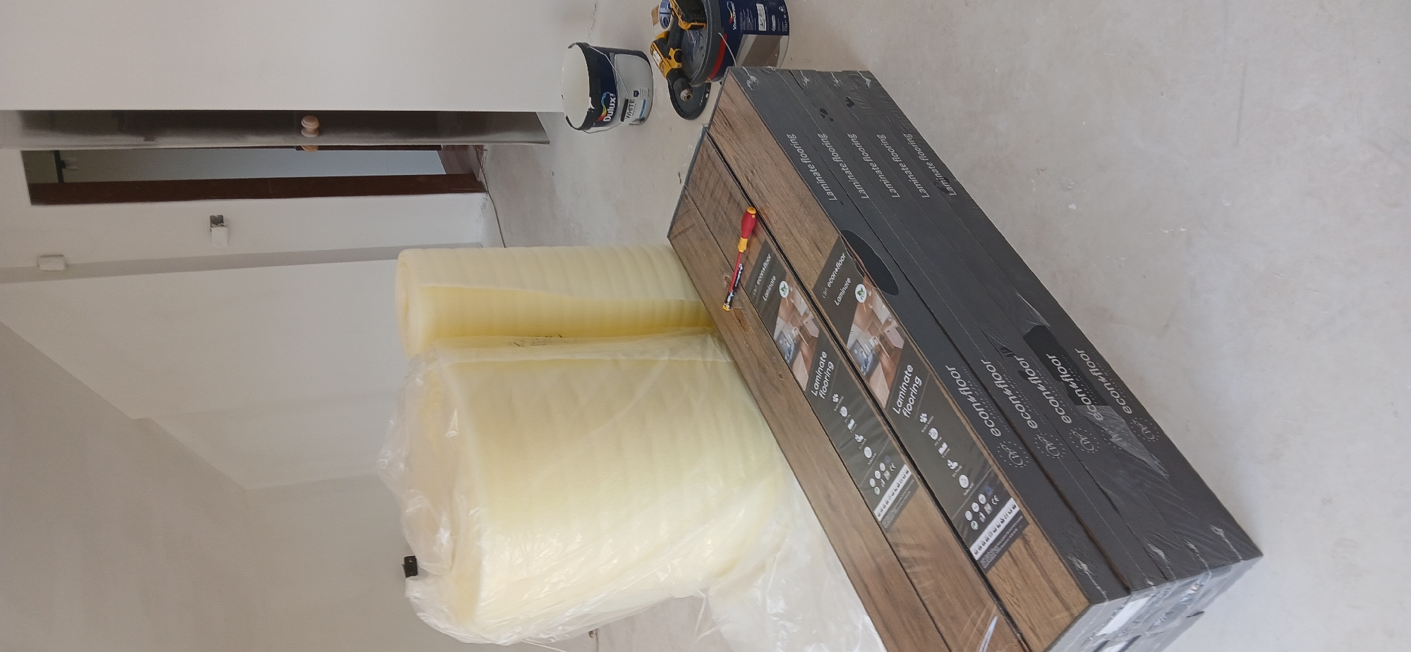 The image shows construction materials in what appears to be an indoor space, likely in Sofia, Bulgaria. Three packs of laminate flooring labeled "econ floor Laminate flooring" are stacked, alongside two rolls of underlayment. A screwdriver rests on top of the flooring packs. In the background, there's a white paint can labeled "Dulux VWHITE," a drill, and a partial view of a door or window frame. The walls are white, suggesting the space is being renovated or prepared for flooring installation.
