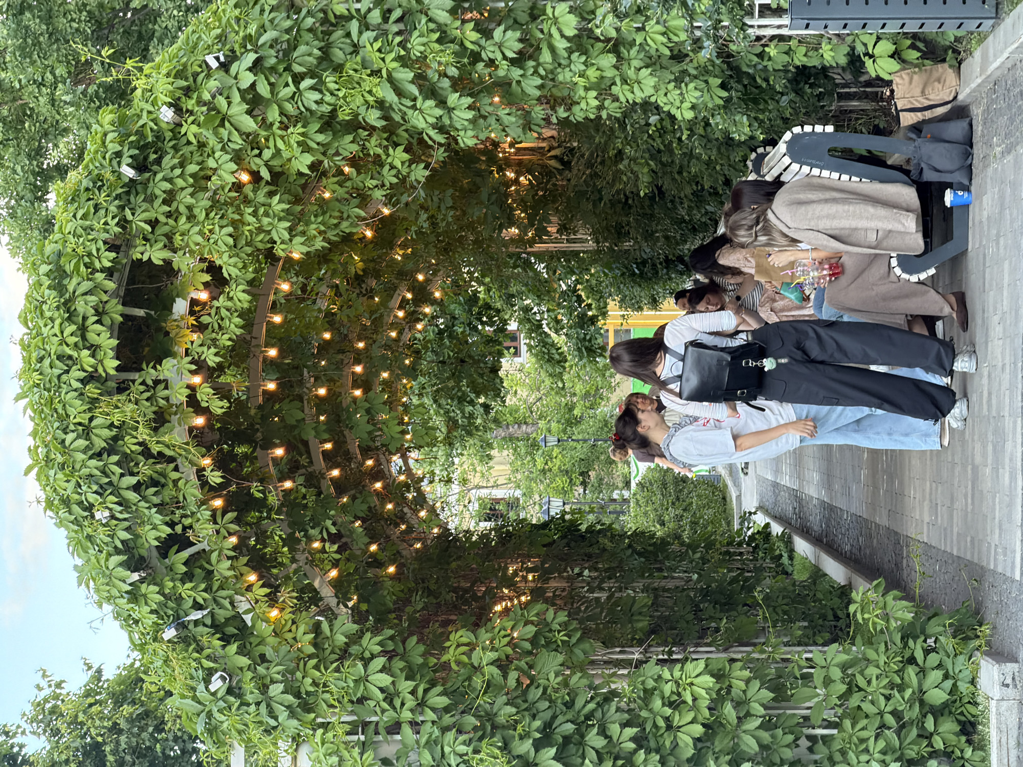 The image shows a group of women standing under an arched trellis covered with green vines and strung with small, warm-toned lights. The women appear to be interacting, some holding drinks. The setting is outdoors on a paved area. The location is identified as Moscow, Russia. The environment suggests an urban park or public space. Details include clothing styles and personal accessories. The general atmosphere seems relaxed and social.