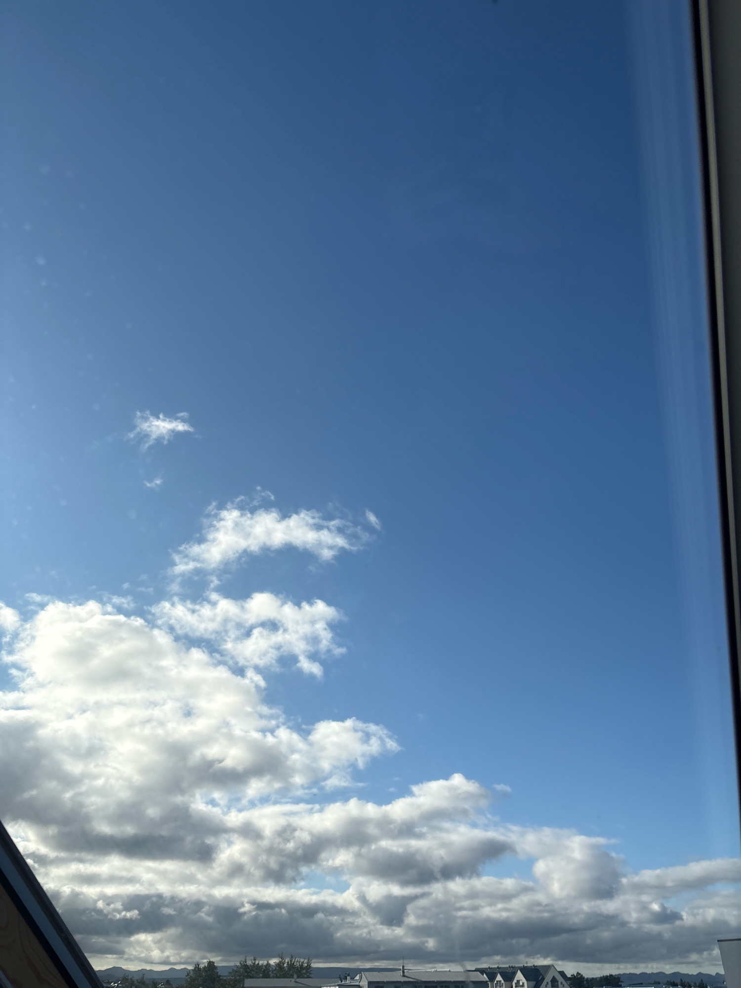 A photograph captures a clear blue sky with scattered white clouds. In the foreground, the edge of a window frame is visible on the right side of the image, and another angled wooden frame is visible on the bottom left. In the distance, a low-lying urban landscape can be seen beneath the clouds, featuring rows of buildings with peaked roofs and trees. The overall impression is of a daytime view from an elevated position in Reykjavik, Iceland, overlooking a residential or commercial area.
