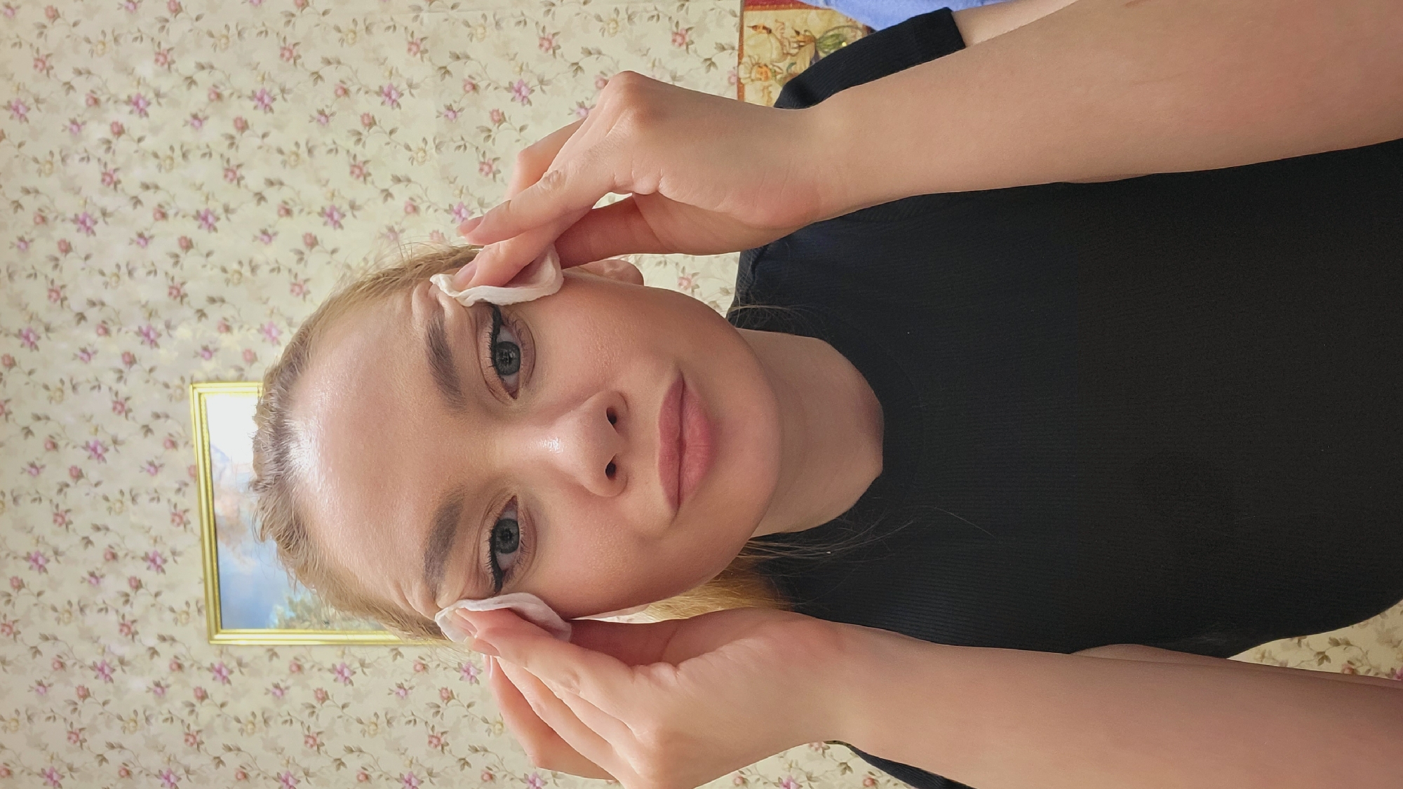 A woman is removing makeup with cotton pads. She is wearing a black shirt. The background is floral wallpaper. There is a framed picture on the wall. The location is Зипуново, Russia.