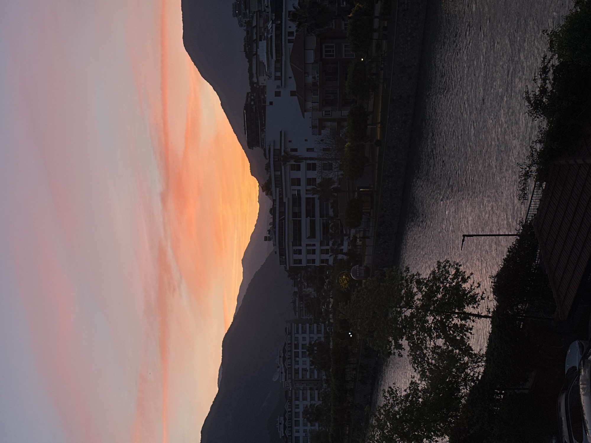 This image captures a serene twilight or dawn scene of a coastal town, likely Alanya, Turkey, as indicated by the location hint. The photograph is oriented vertically but depicts a horizontal landscape.
The upper left portion of the image is dominated by a dramatic sky, painted with soft gradients of pale blue, delicate pink, and vibrant orange-yellow clouds. This colorful sky suggests either a beautiful sunrise or a tranquil sunset.
Below the colorful sky, a prominent, dark mountain range stretches across the mid-ground, silhouetted against the brighter light of the horizon. Its rugged peaks frame the urban landscape below.
Clustered along the base of the mountains and extending towards the waterfront are numerous multi-story buildings, appearing as dark silhouettes against the sky or faintly lit by the ambient light. These structures appear to be modern residential or commercial buildings, featuring multiple windows and balconies, characteristic of a developed coastal city.
The right side of the image is occupied by a calm body of water, likely the sea or a bay, reflecting the subdued light of the sky. Its surface shows subtle ripples, indicating a gentle breeze or current. Along the edge of the water and interspersed among the buildings, various trees and green foliage are visible, adding natural elements to the urban setting. A streetlight pole is visible near the water's edge, and a car is partially visible in the bottom right foreground, indicating human presence and infrastructure.
No specific activity or event is taking place; the scene conveys a peaceful moment at the transition between day and night. There is no visible text in the image.