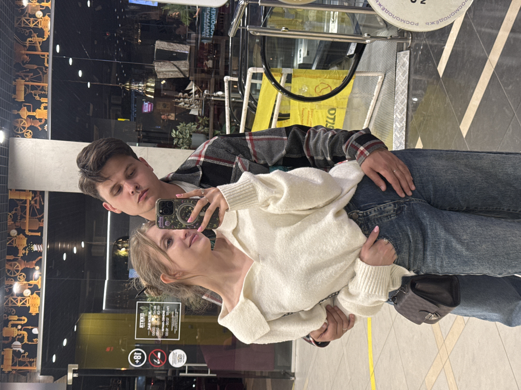 A young couple, a male and a female, are taking a selfie indoors. The man has short brown hair and is wearing a gray and black plaid shirt over a white t-shirt, and blue jeans. He has a ring on his left ring finger. The woman has blond hair, a white sweater, and blue jeans, and is holding a cell phone, using it to take the selfie. She is wearing rings on her fingers and has a small black purse on her left hip. They are standing in a commercial space with black and white floor tiles and store windows. The environment includes various signs, including an "18+" sign and others with Russian text, as well as decorative elements on the walls. There's also a partial reflection of an escalator and other elements of the surrounding infrastructure.