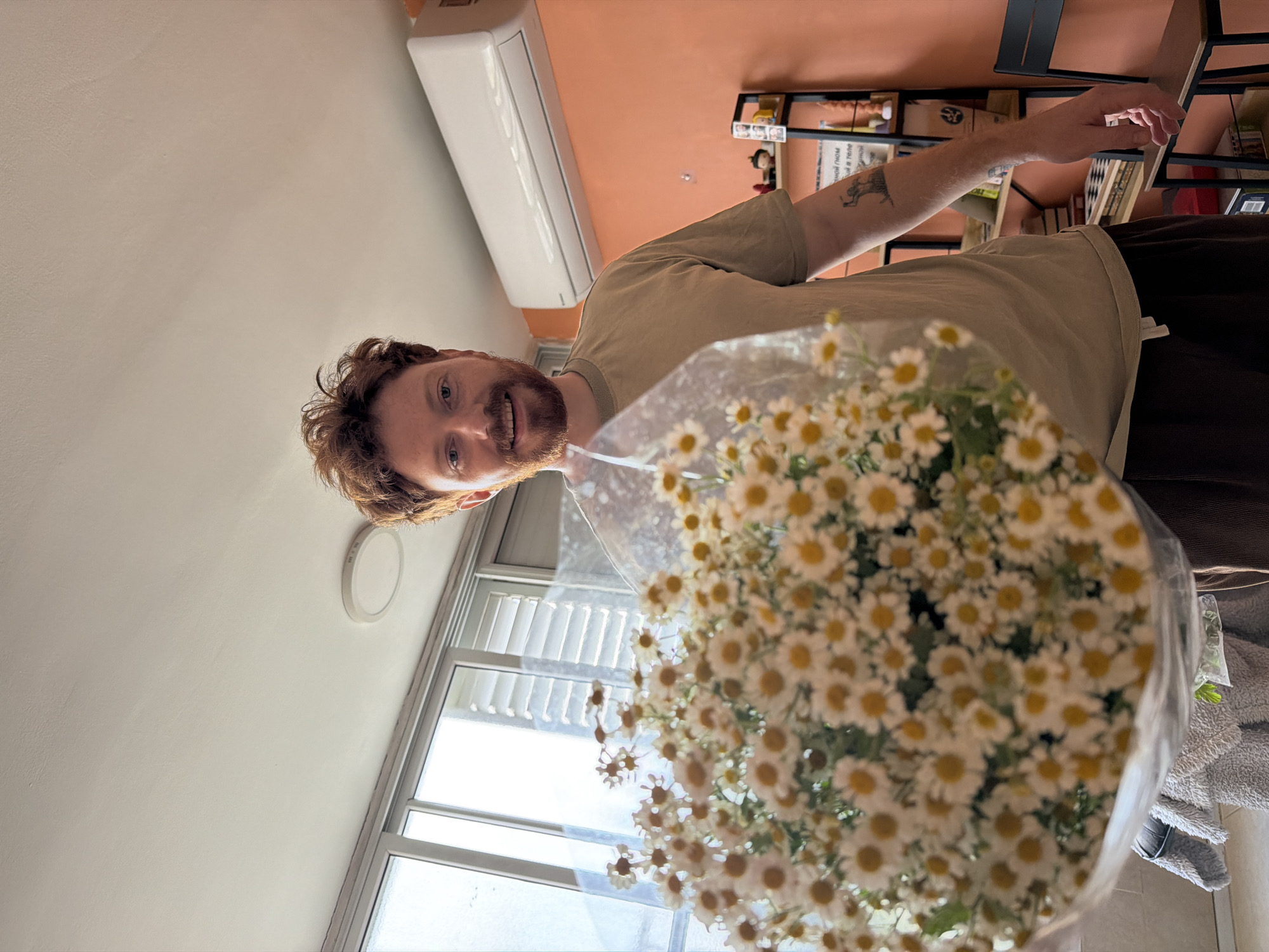 A man with red hair and a beard is holding a large bouquet of daisies in front of him. He is wearing a beige t-shirt and dark pants. Behind him is a window, and a bookshelf with books is visible in the background. An air conditioning unit is mounted on the wall. Based on the architectural style and general environment, the location is likely Tel-Aviv, Israel.