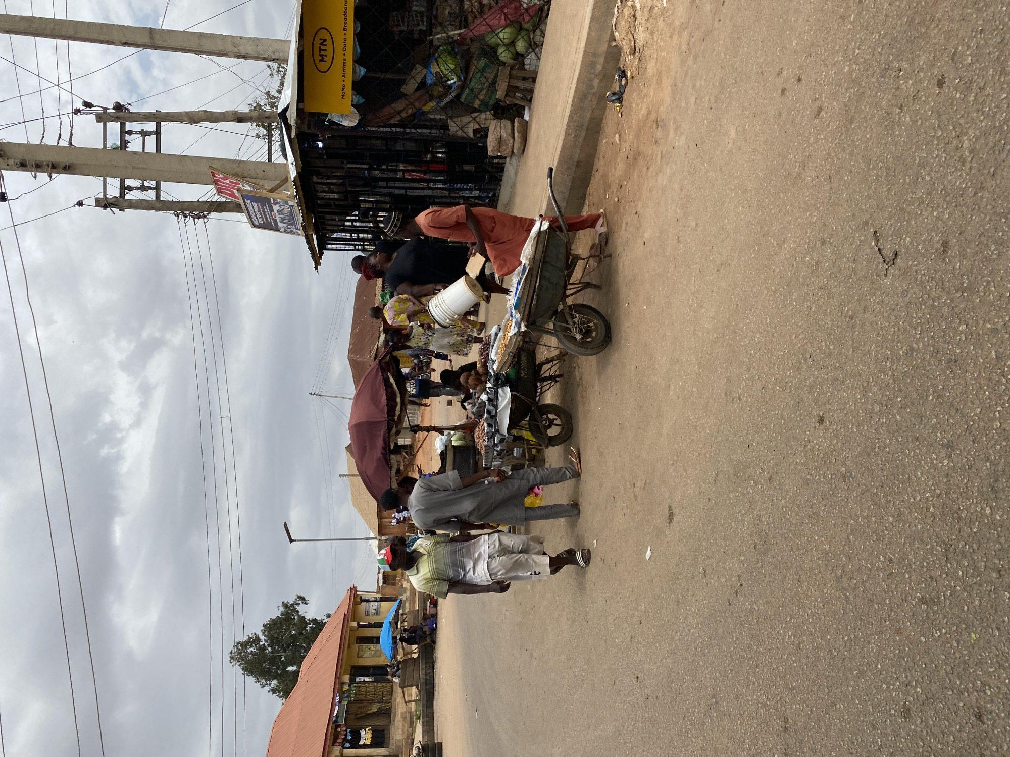 The image shows a street scene in Bukuru, Nigeria. There is a vendor selling goods from a wheelbarrow. People are gathered around the vendor. There are buildings on either side of the street. There is a sign for MTN. Power lines are visible above the scene. The sky is overcast.