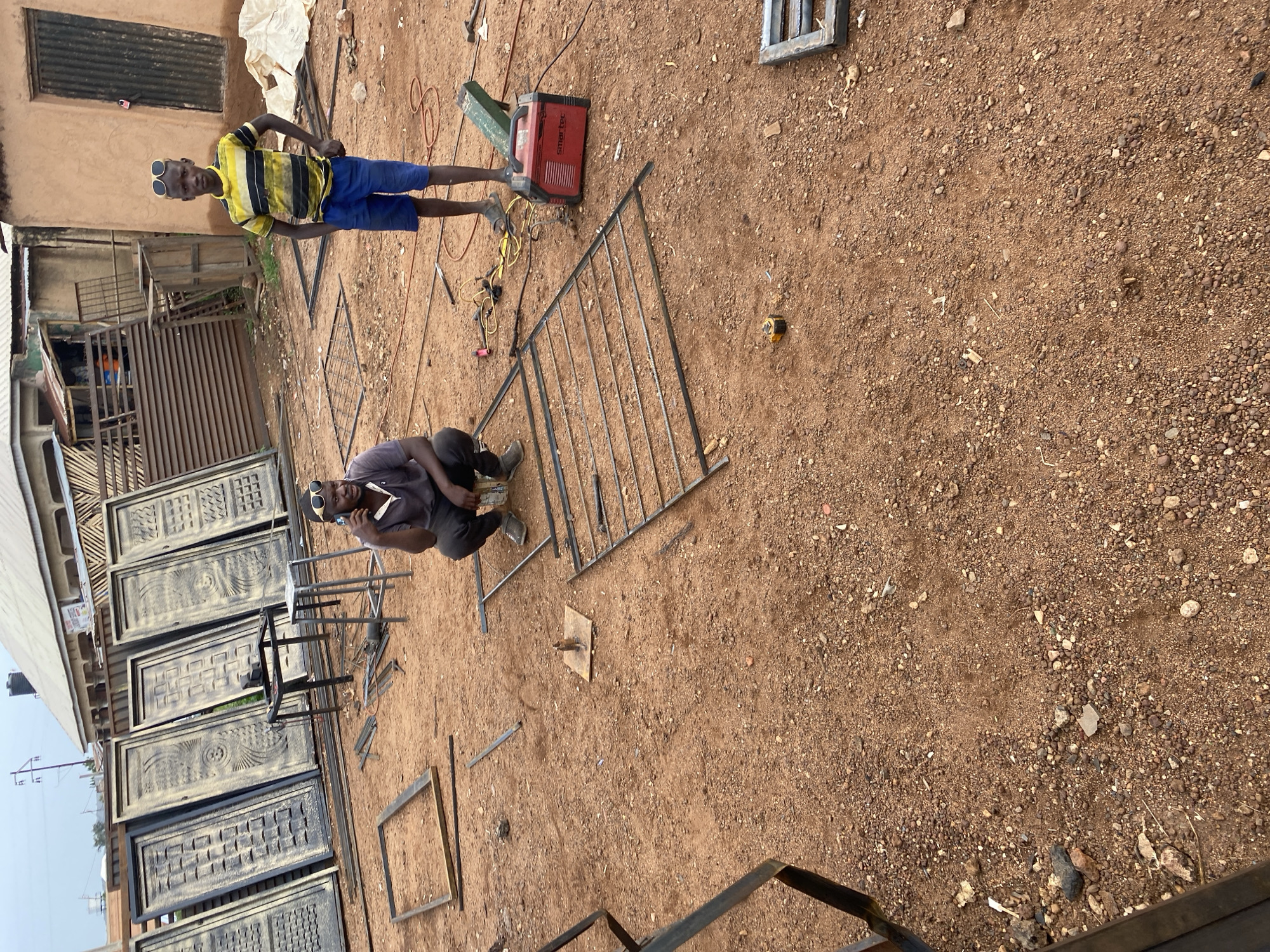 The image shows an outdoor workspace in Bukuru, Nigeria. Two men are present; one is standing wearing sunglasses, a yellow and black striped shirt, and blue shorts. The other is kneeling, also wearing sunglasses and a grey shirt. Scattered around them are metal gates and door panels, some finished and painted, others in the process of construction. A red "Smartec" welding machine is visible near the standing man. The ground is dirt and gravel. Buildings are visible in the background.