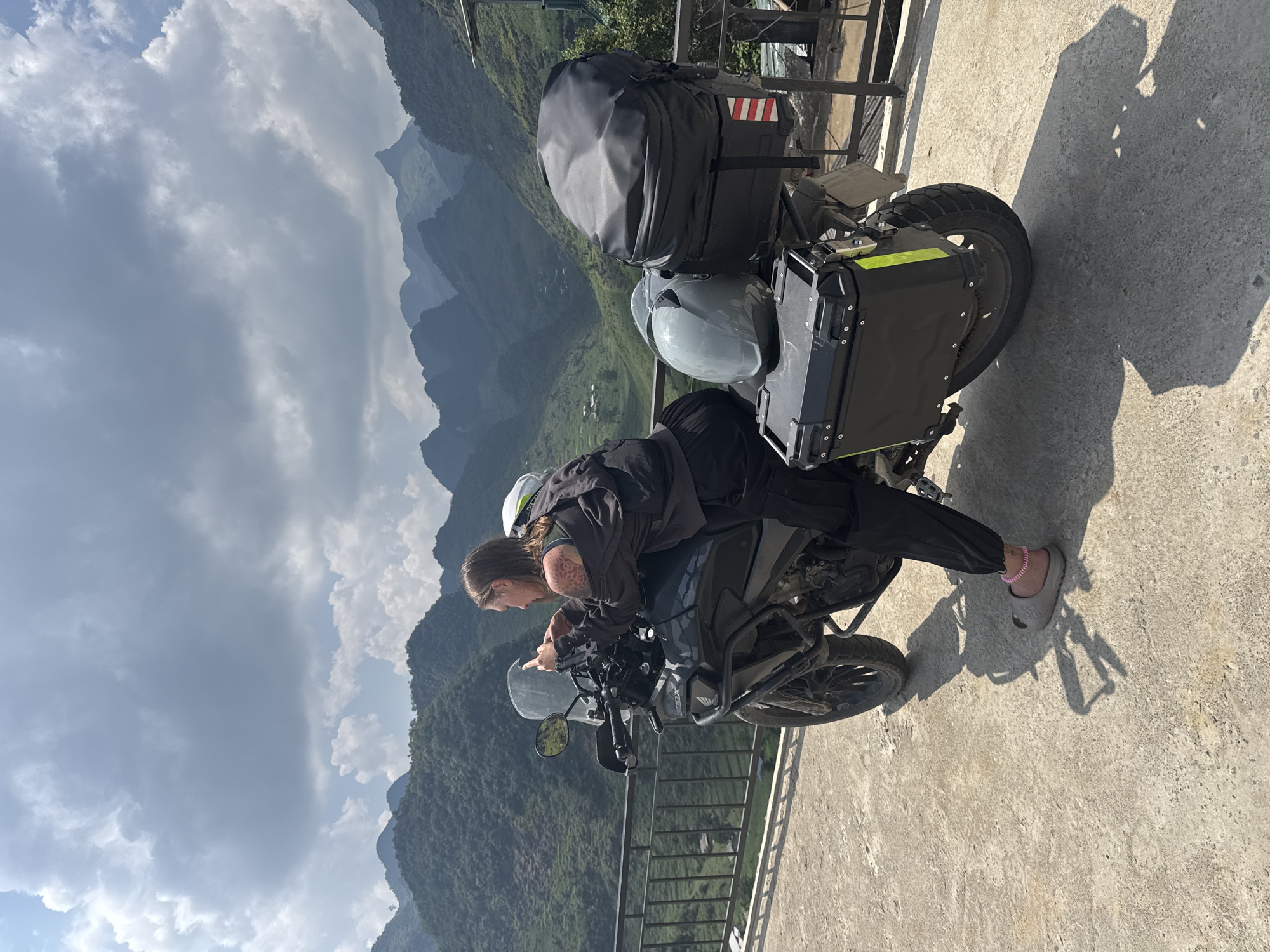 A traveler in dark clothing and sandals is leaning over a motorcycle parked near a metal railing. The motorcycle has luggage, including a large black bag, a gray helmet, and a black side case with a reflective stripe. Behind the motorcycle and rider, there is a mountainous landscape covered in vegetation under a cloudy sky. The photo was taken in Lũng Phìn Commune, Vietnam. The surface beneath the motorcycle appears to be concrete.