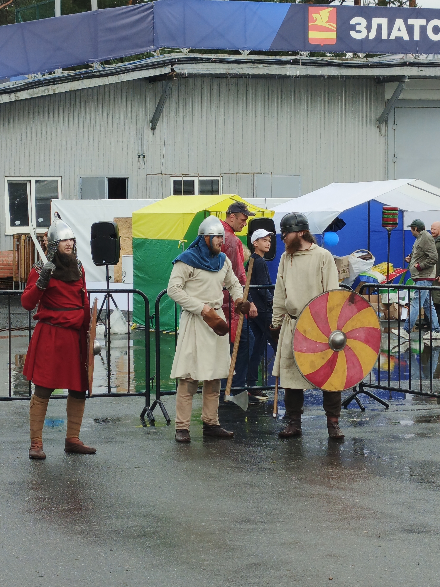 The image depicts an outdoor event in Zlatoust, Russia, featuring individuals dressed in historical reenactment attire. In the foreground, two men stand in medieval-style costumes. The man on the left wears a red tunic, chainmail, a metal helmet, and leg wraps, holding a sword and a round shield. The man on the right is dressed in a light-colored tunic, a helmet, and leg coverings, carrying a shield with a red and yellow radial pattern and an axe. Behind them, other individuals are visible, some in casual wear and others also in costume, near tents and a building. The ground is wet, suggesting recent rain, and reflections are visible on the asphalt. A banner with the text "ЗЛАТОУСТ" and a coat of arms is displayed on the building in the background. The overall atmosphere suggests a public gathering or festival.